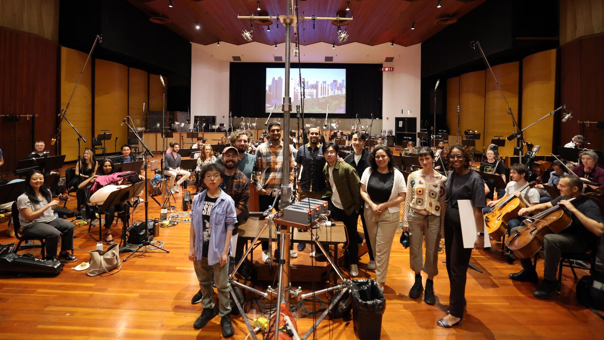 Group of diverse people pose in a large rehearsal hall; an orchestra sits with instruments in the background; microphone stands and recording gear fill the room and a city skyline appears on a screen.
