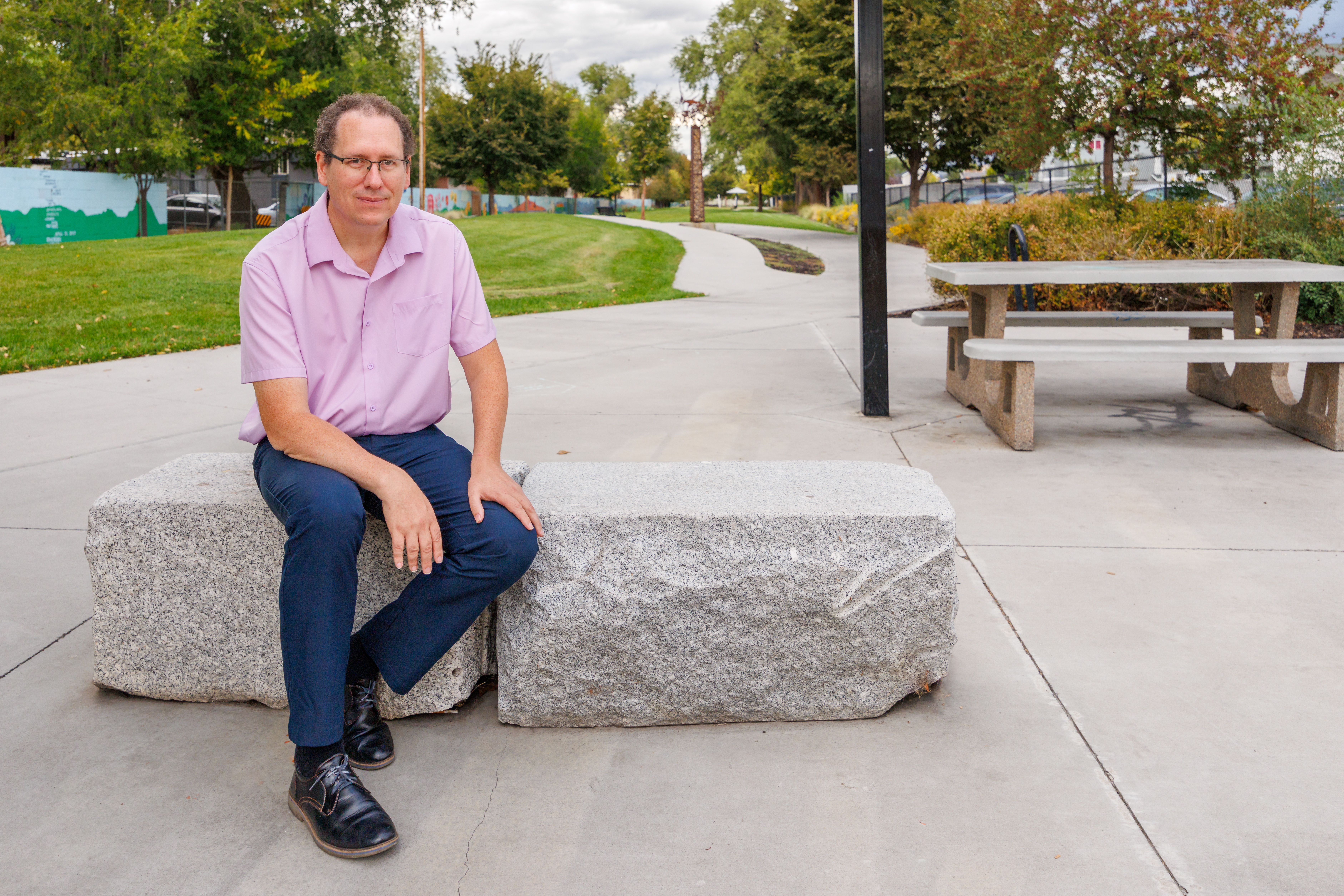 Man in a pink shirt and blue pants sitting on a large stone bench in an outdoor park with trees, concrete path, and picnic table nearby under a cloudy sky.