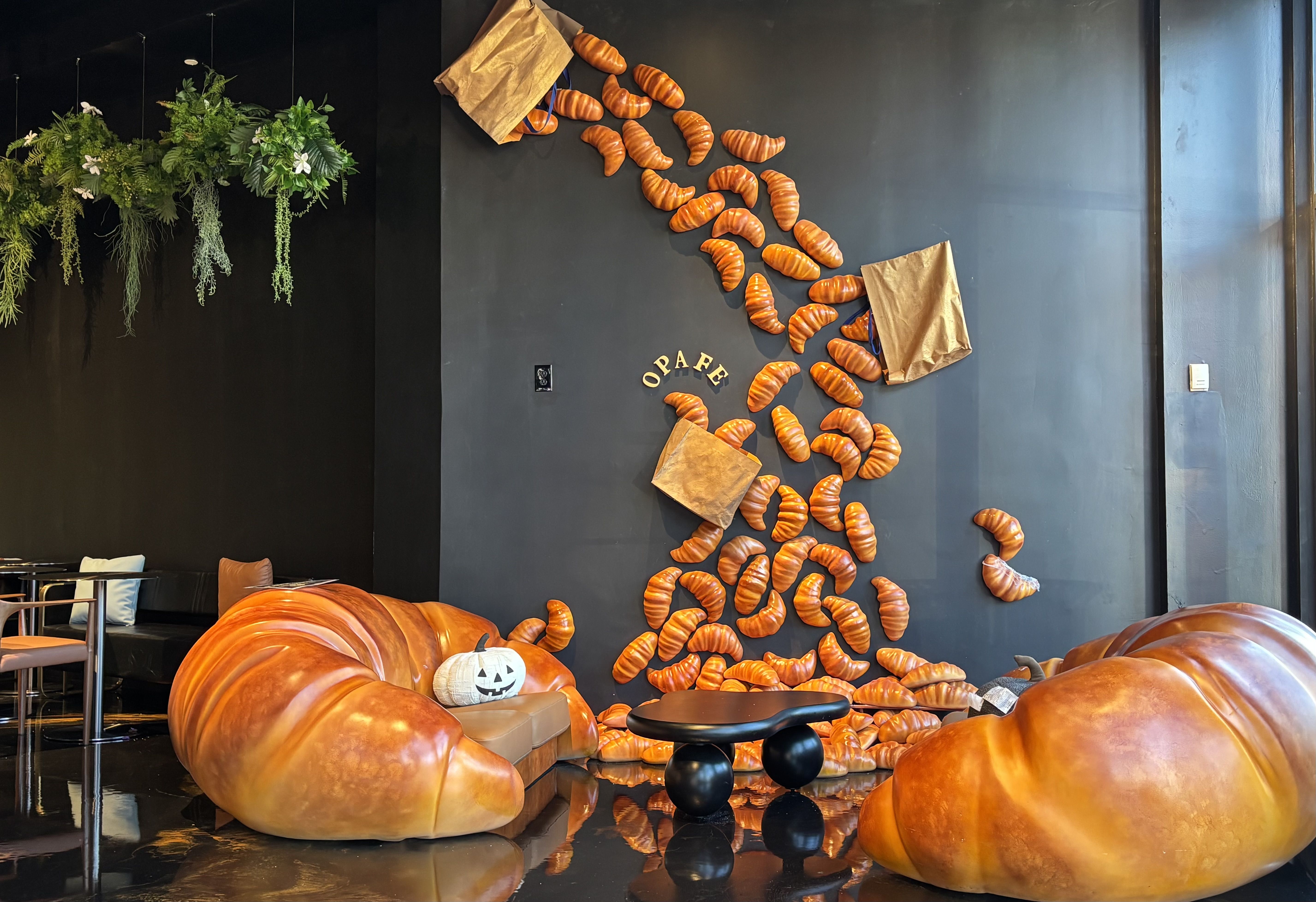 Cafe interior with dark wall and orange croissant-shaped pieces cascading down; green hanging plants on left; brown paper bags; pumpkin cushions; small white jack-o'-lantern on floor.