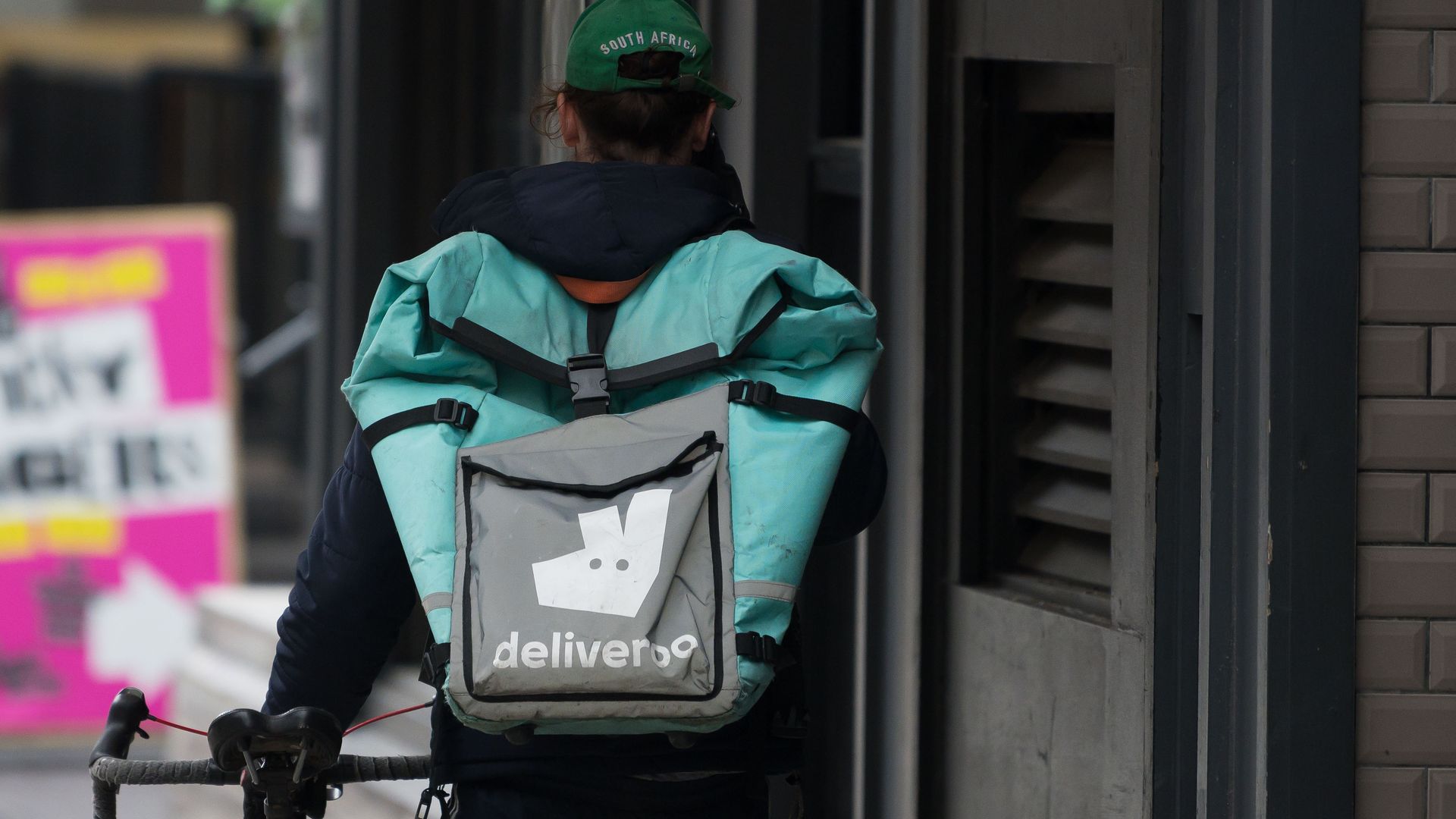 In this image, a Deliveroo deliveryperson walks down the street with a package on their back and guiding a bicycle.