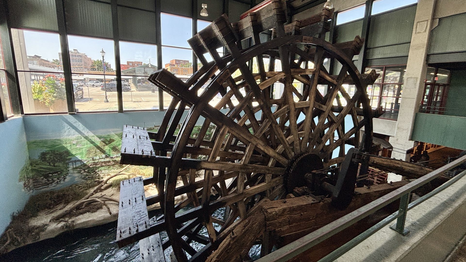 Inside a modern atrium, a large wooden water wheel dominates the scene. Big windows reveal a street with cars outside, while a river mural runs along the blue lower wall.