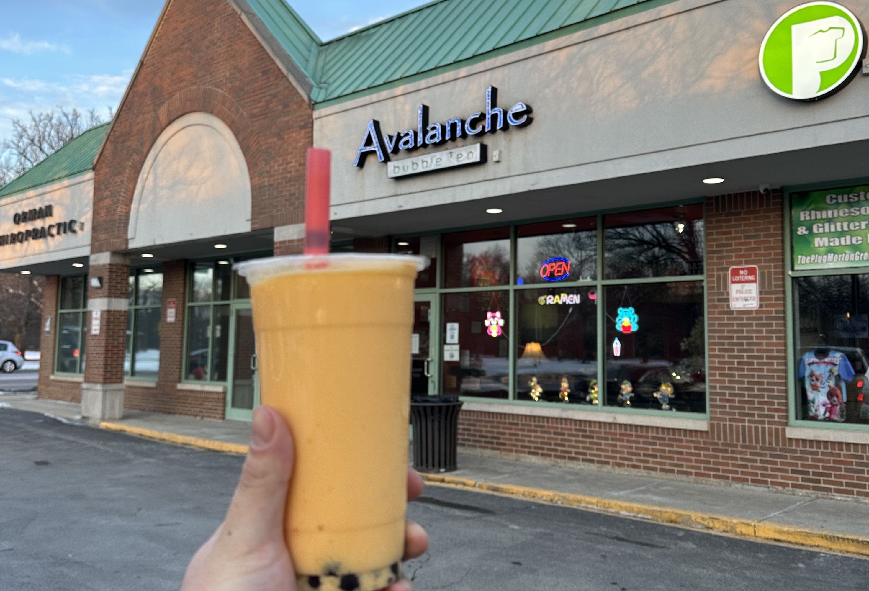 A hand holding an orange bubble tea with black tapioca pearls, in front of a brick storefront reading "Avalanche bubble Tea" with a neon open sign in the window.