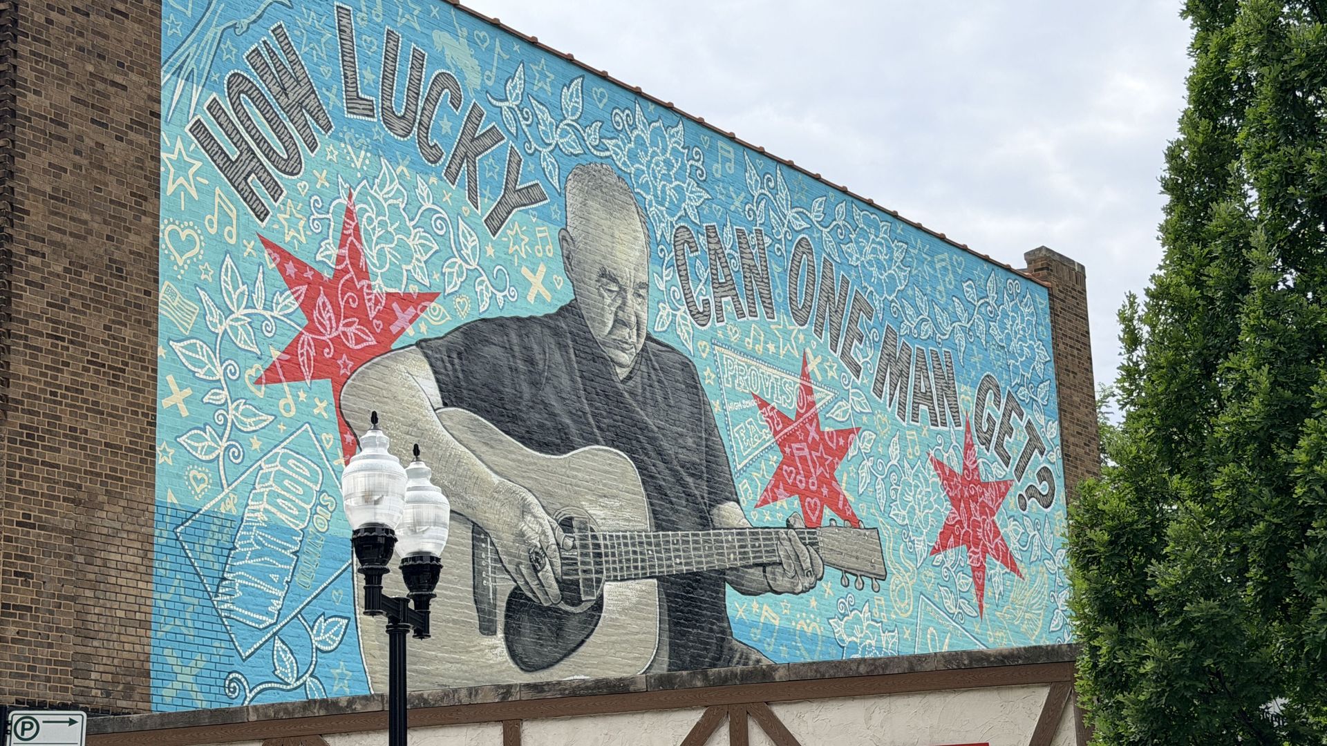 Large mural on a brick wall featuring a grayscale guitarist with a guitar, set against a blue decorative background with stars, leaves, and musical notes; trees to the right and a cloudy sky.