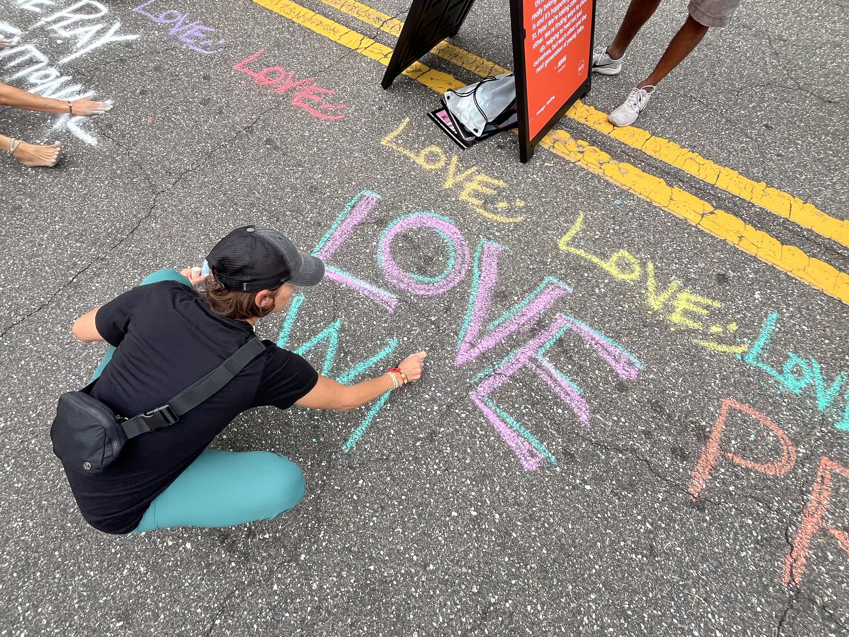 People kneeling on a cracked asphalt surface, drawing colorful chalk art. Bright pink, teal, and purple letters surround yellow painted lines. A black bag and an orange information sign sit nearby.