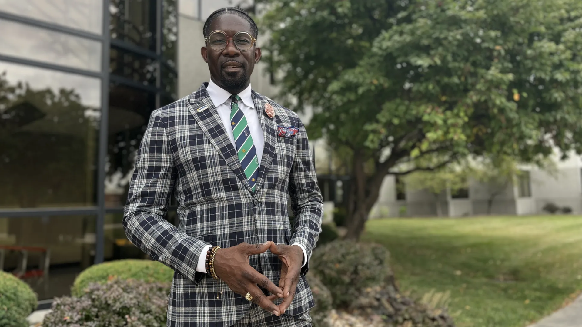 DMPS Superintendent Ian Roberts wearing a black and white plaid suit with a green striped tie and round glasses, standing outdoors near bushes and a building with glass windows.