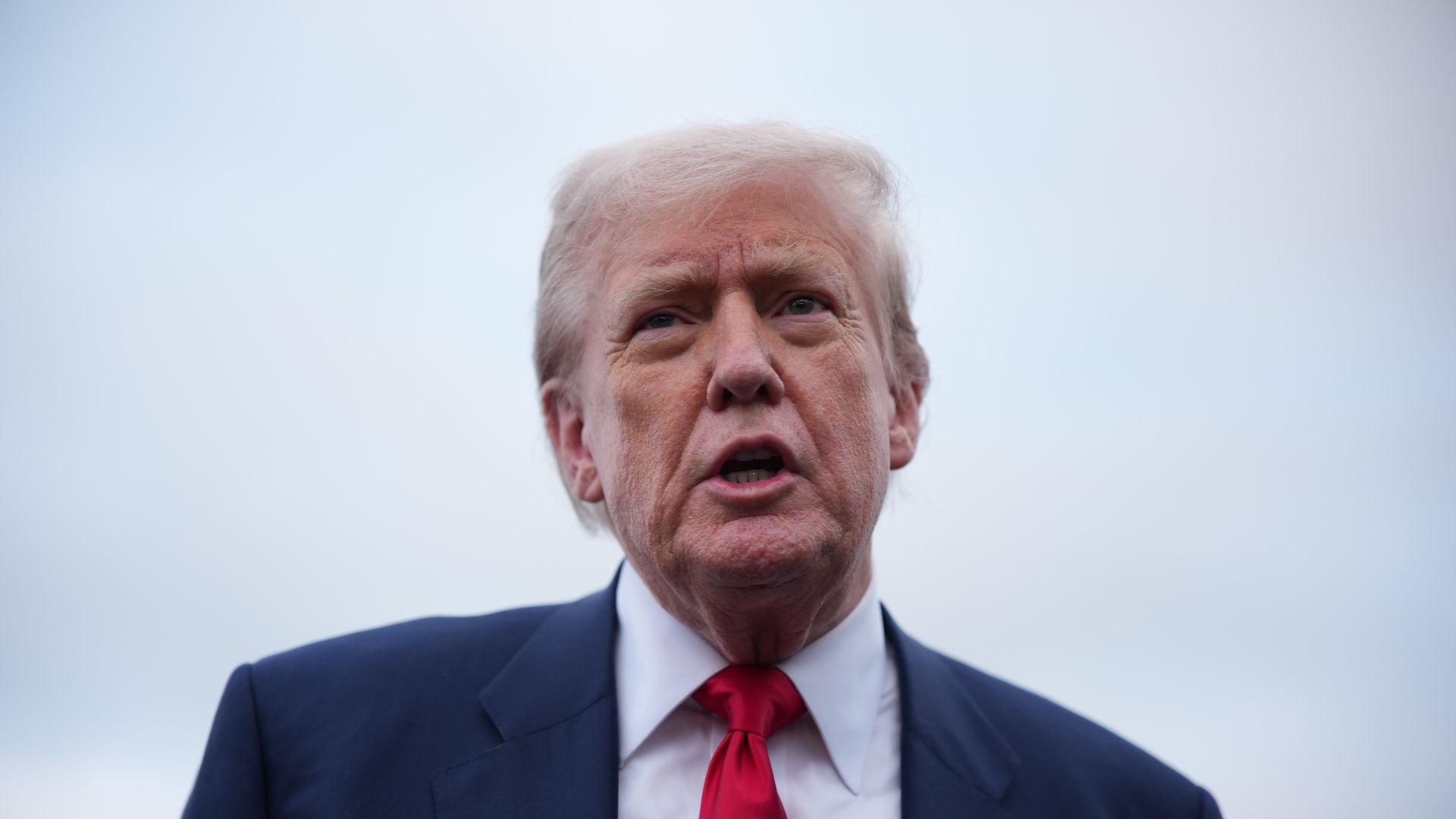 Close-up of a man in a navy suit, white shirt, and red tie with an American flag pin, speaking outdoors against a cloudy sky.