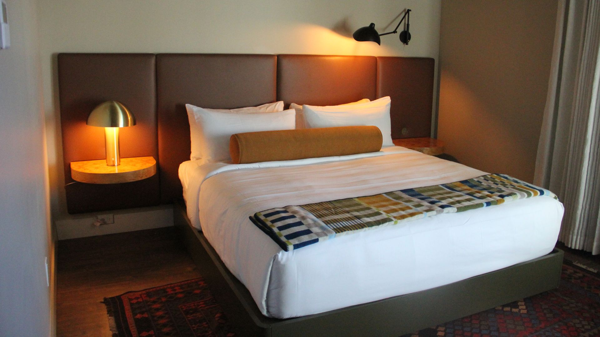 Modern hotel room with a king bed, white linens, and a long brown bolster. A padded headboard spans the wall with lamps on each side and a plaid throw at the foot.