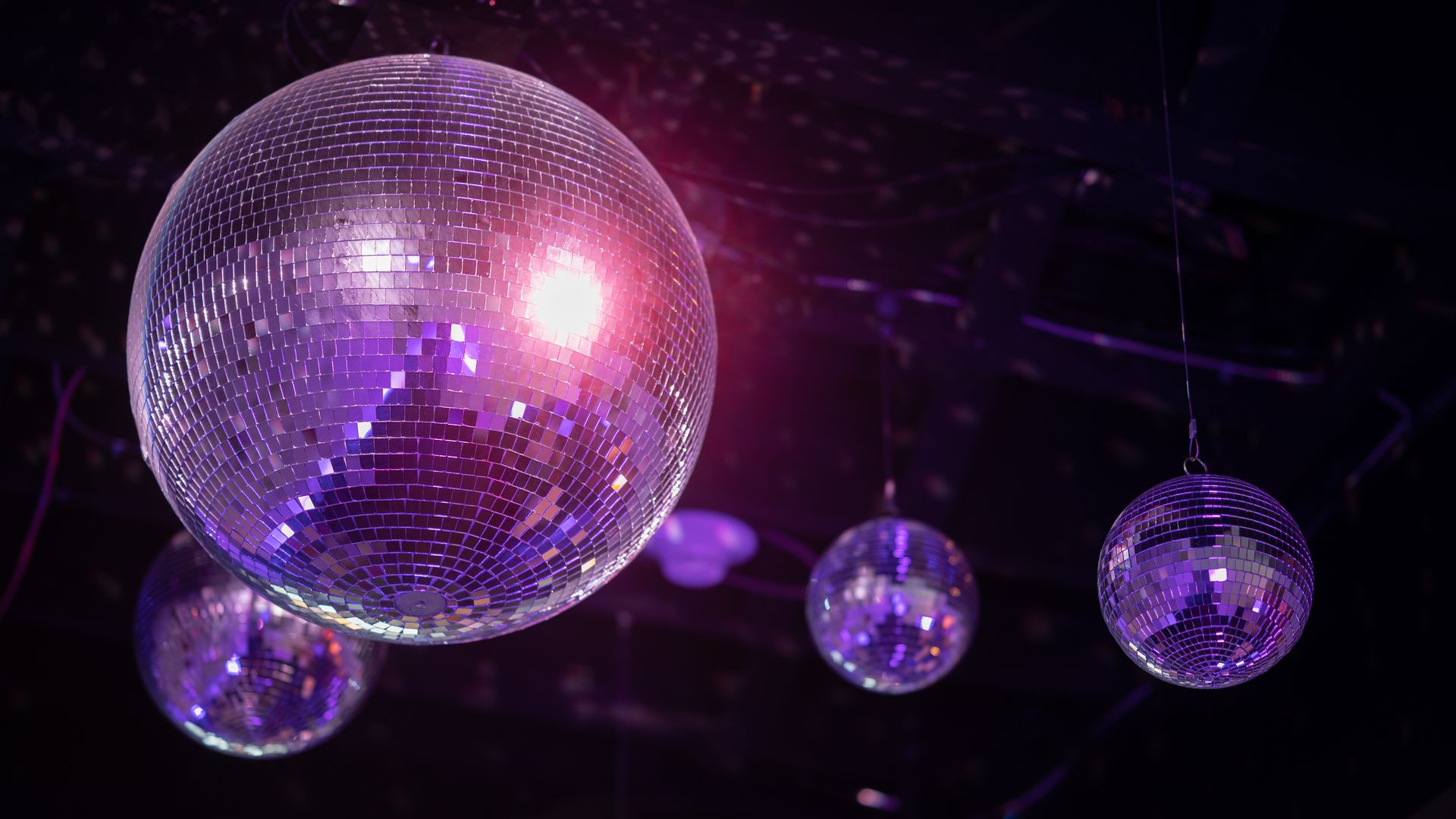 Four mirror disco balls of varying sizes hanging from a dark ceiling, reflecting purple and pink light in a dimly lit party or club setting.