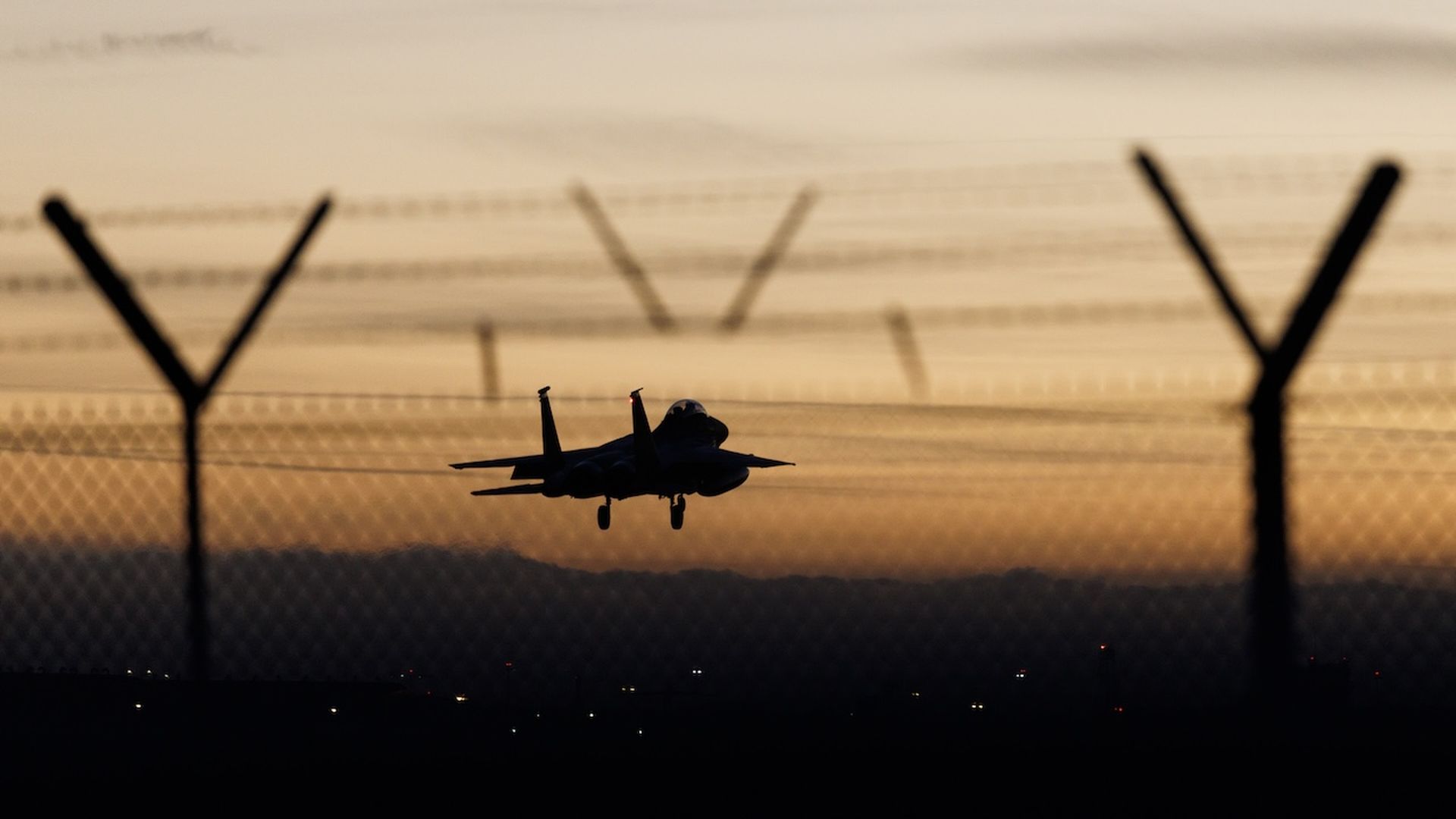 Silhouette of a jet fighter taking off with landing gear down against an orange sunset, framed by a barbed wire fence and dark landscape.