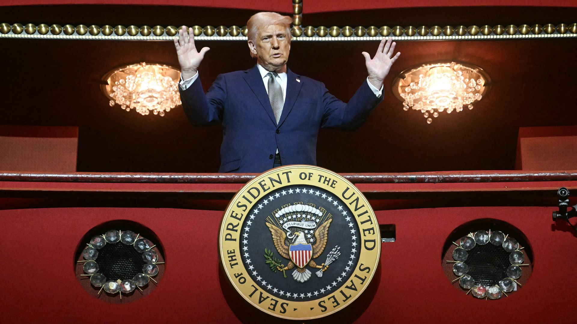 President Trump, wearing a navy jacket with a US flag pin at the top of his left lapel, white shirt and silver tie, holds both hands aloft while standing in the red  presidential box at the John F. Kennedy Center for the Performing Arts.