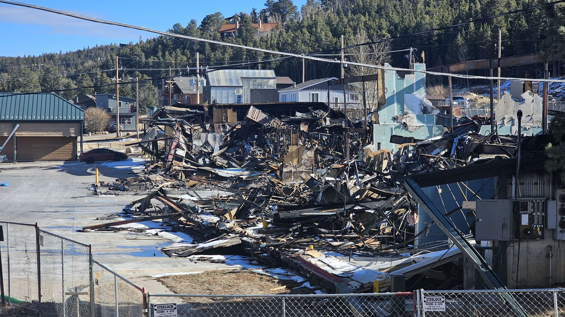 Ruined building with collapsed walls and rubble, surrounded by a chain-link fence and parking lot, with green trees and houses in the background under a clear blue sky.
