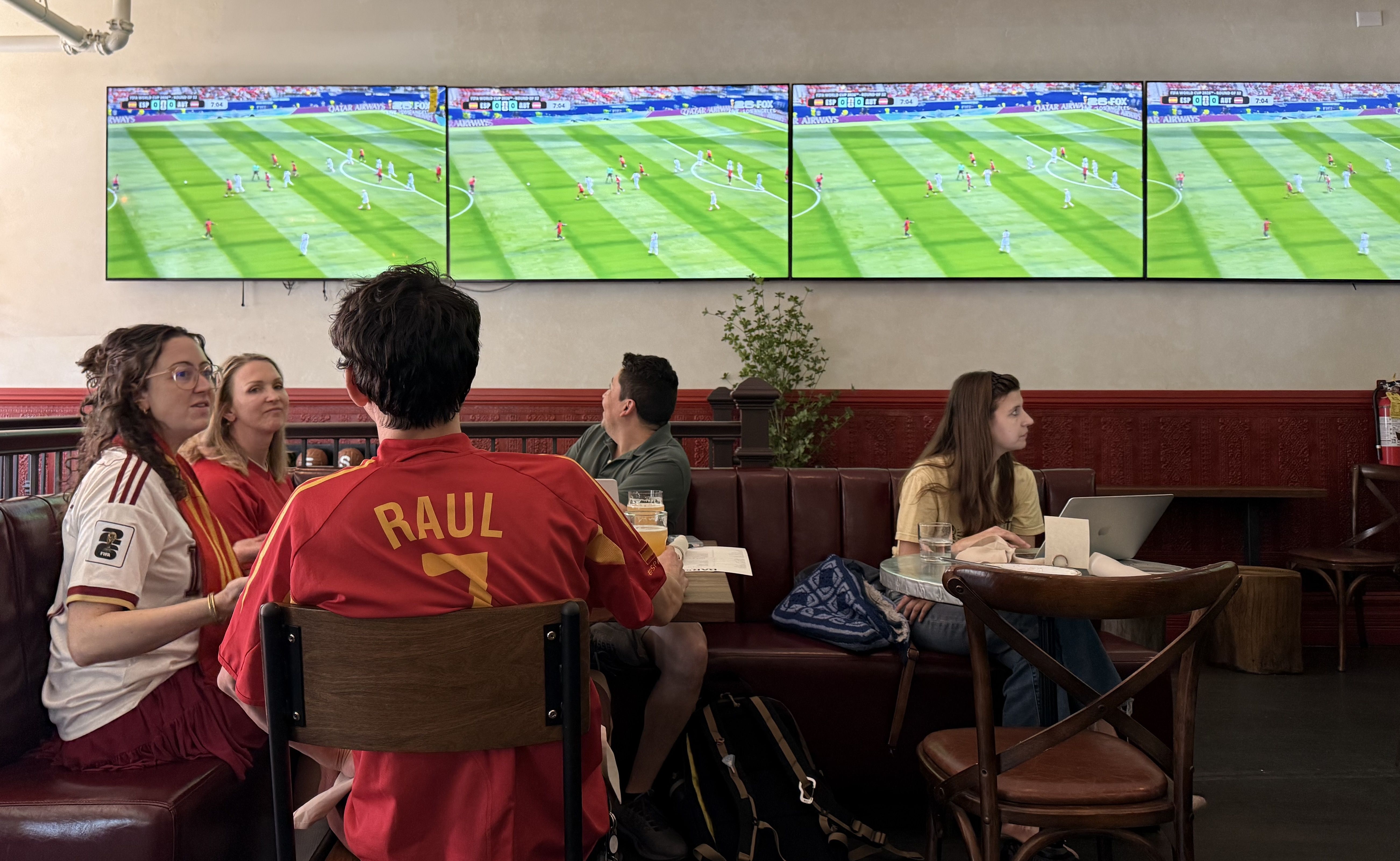 A sports bar with four wall-mounted screens showing a soccer match. Patrons sit at a booth, including a man in a red Raul 7 jersey, watching with drinks and a laptop nearby.