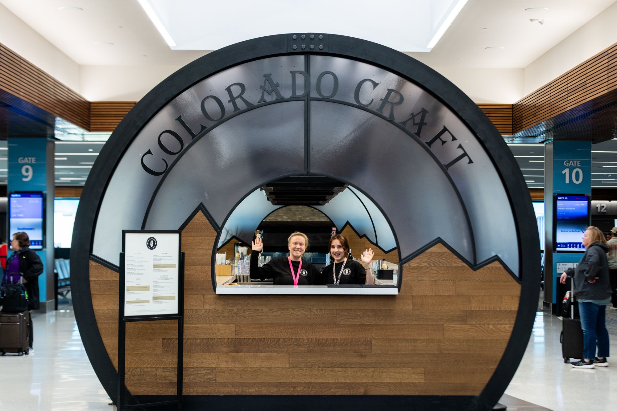 Inside a modern airport, a large circular dark metal arch frames a counter with the sign "COLORADO CRAFT". Two staff wave from behind the bar as Gate 9 and 10 signs and travelers appear nearby.