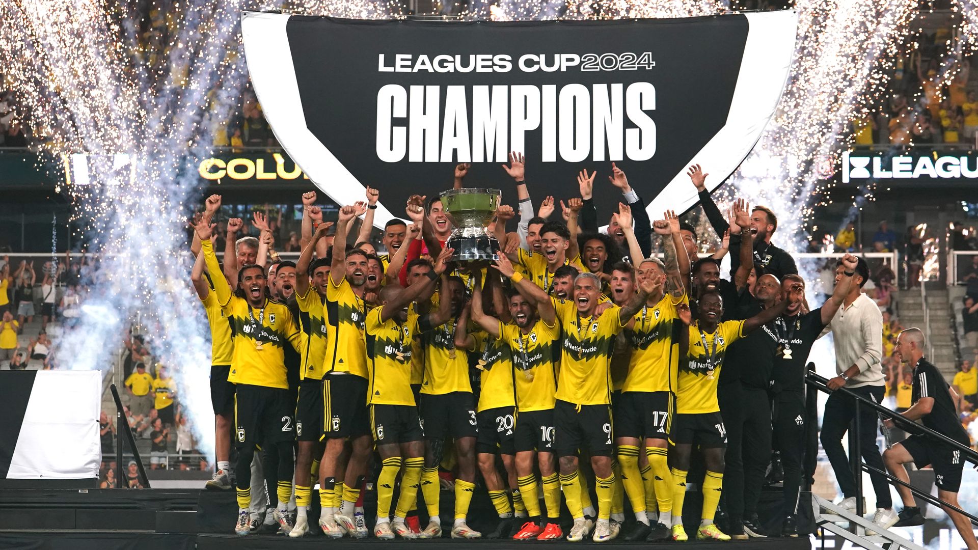 Crew players hoist the Leagues Cup trophy in front of a banner that says "Leagues Cup 2024 Champions" in front of confetti cannons