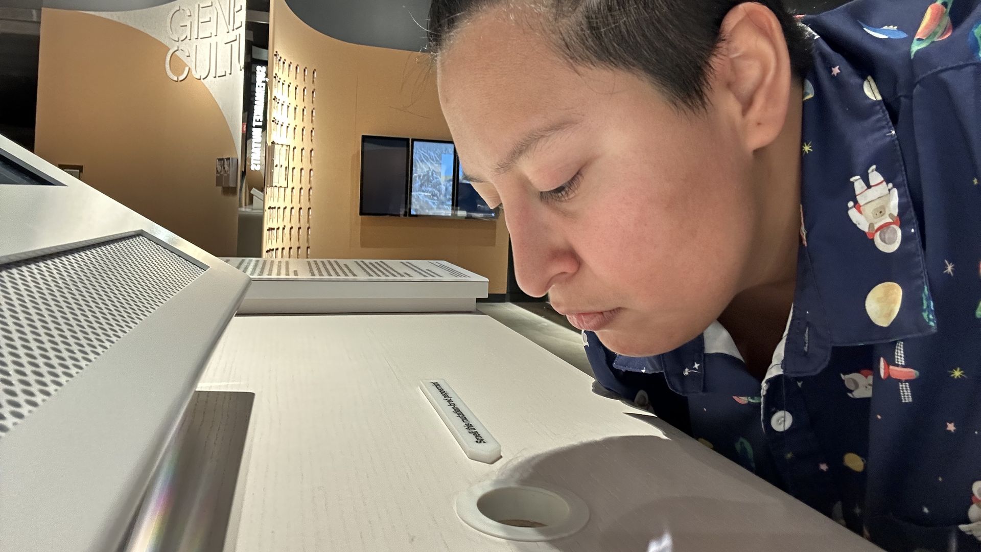 Steph Solis on the right hovers their nose over a hole with a small sign pointing to the hole saying it contains a "mutagenic" peppermint scent.