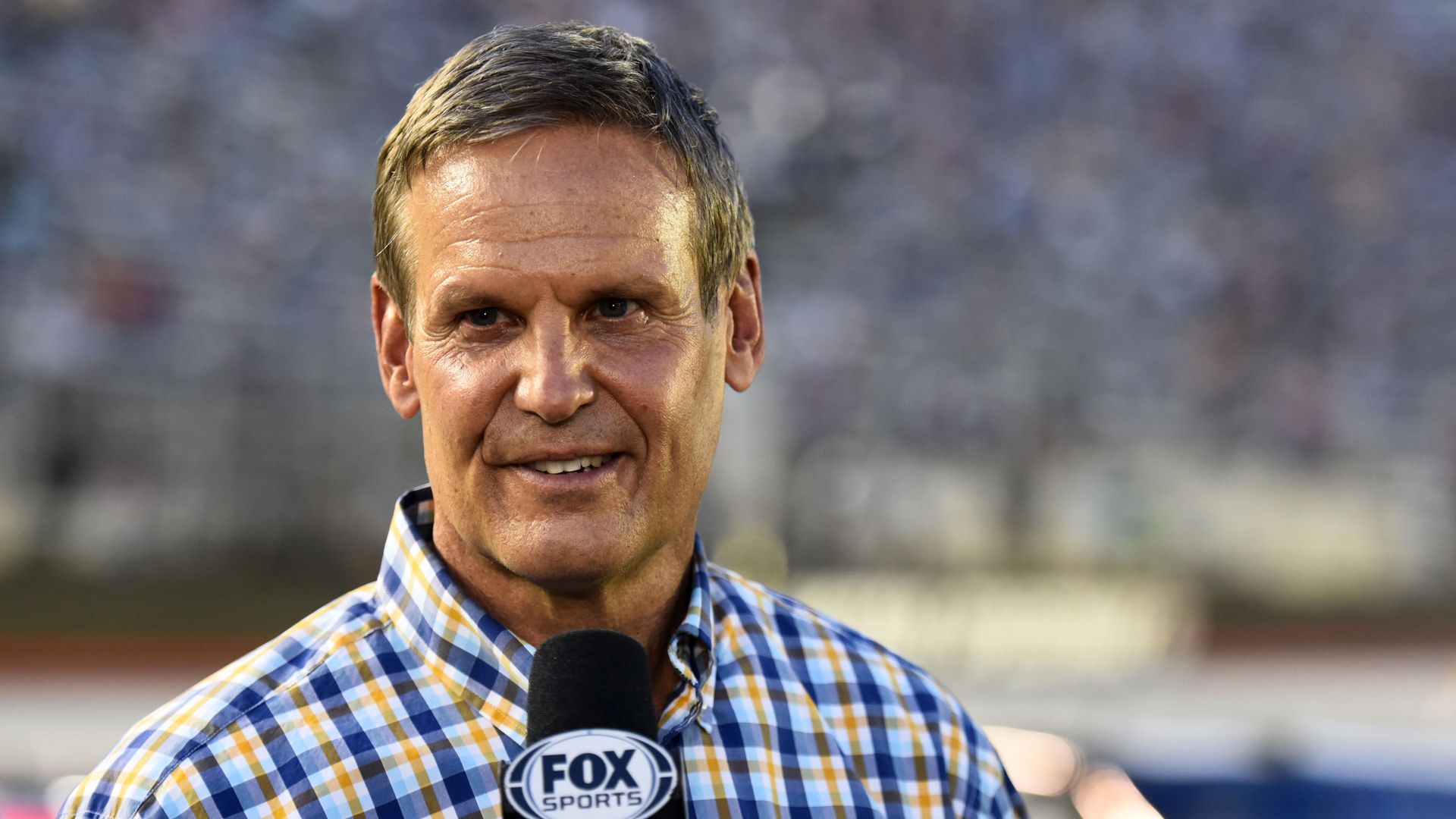 Photo of Bill Lee in a plaid shirt speaking into a Fox microphone at a stadium