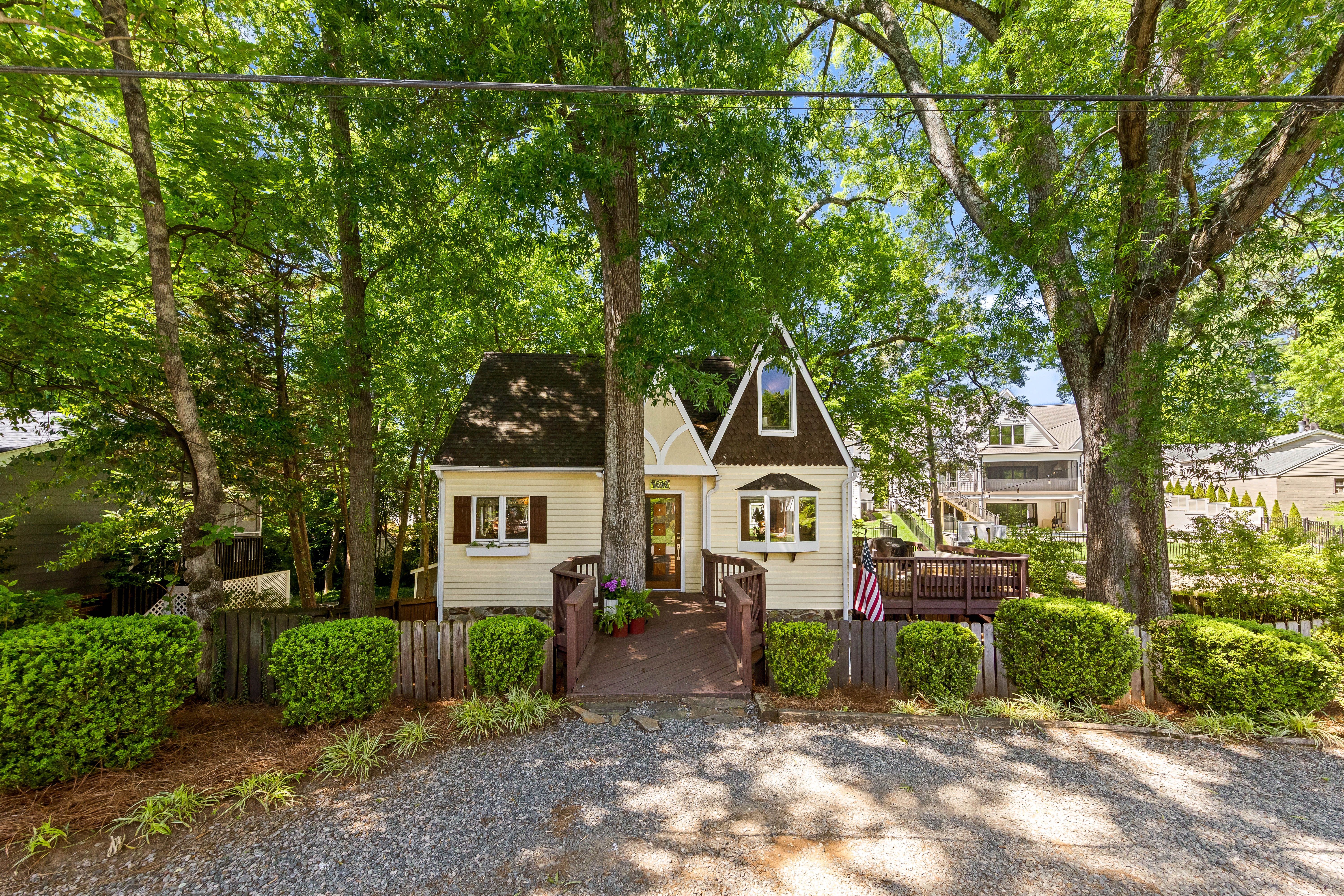 house for sale Cozy cream cottage with a brown gabled roof and white trim, front porch, nestled among dense green trees. Wooden fence, gravel driveway, and an American flag beside neighboring houses.