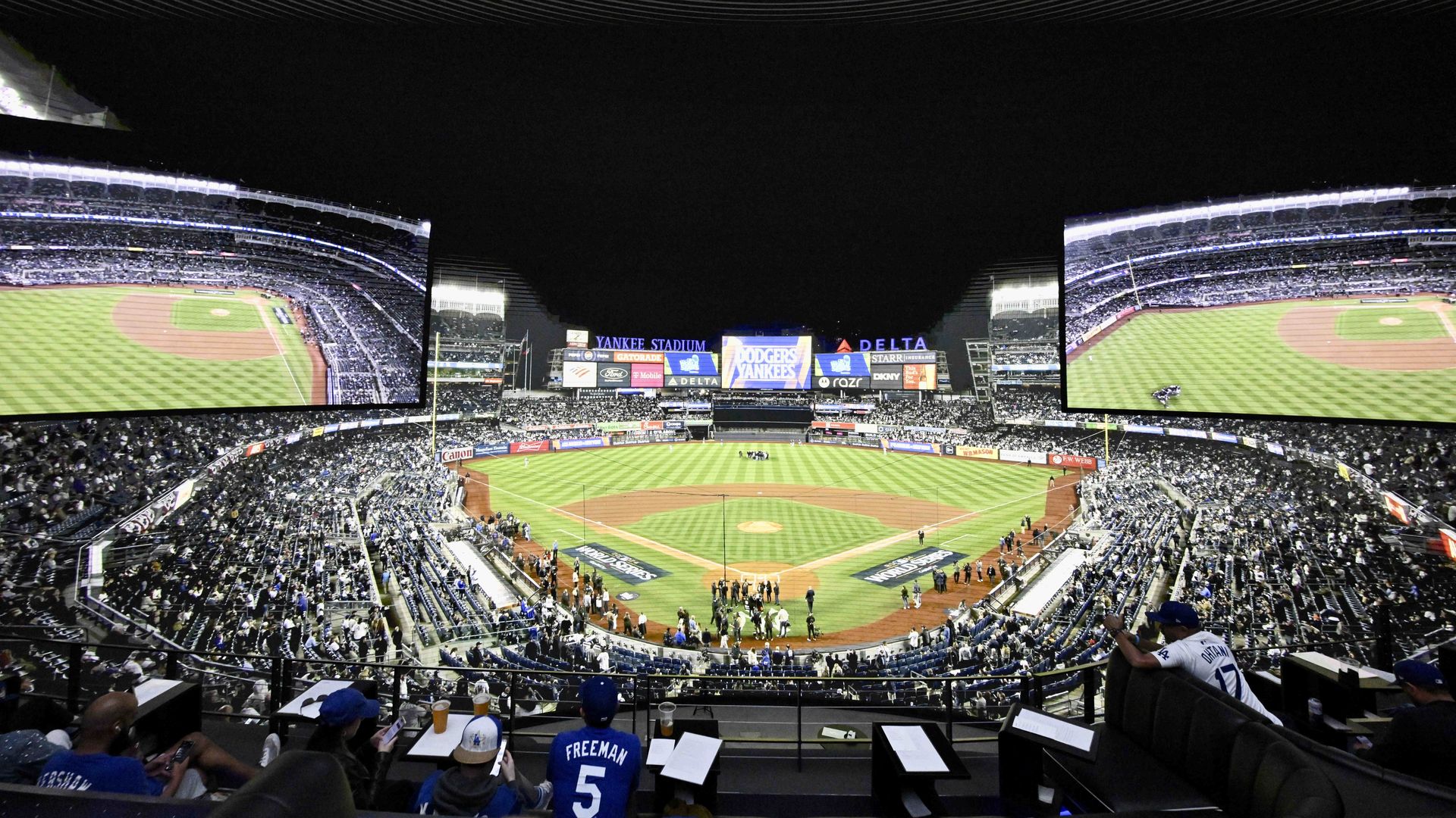 Wide view of Yankee Stadium at night with fans wearing Dodgers gear, large screens showing the baseball field, and scoreboard displaying "Dodgers Yankees" for a game event.