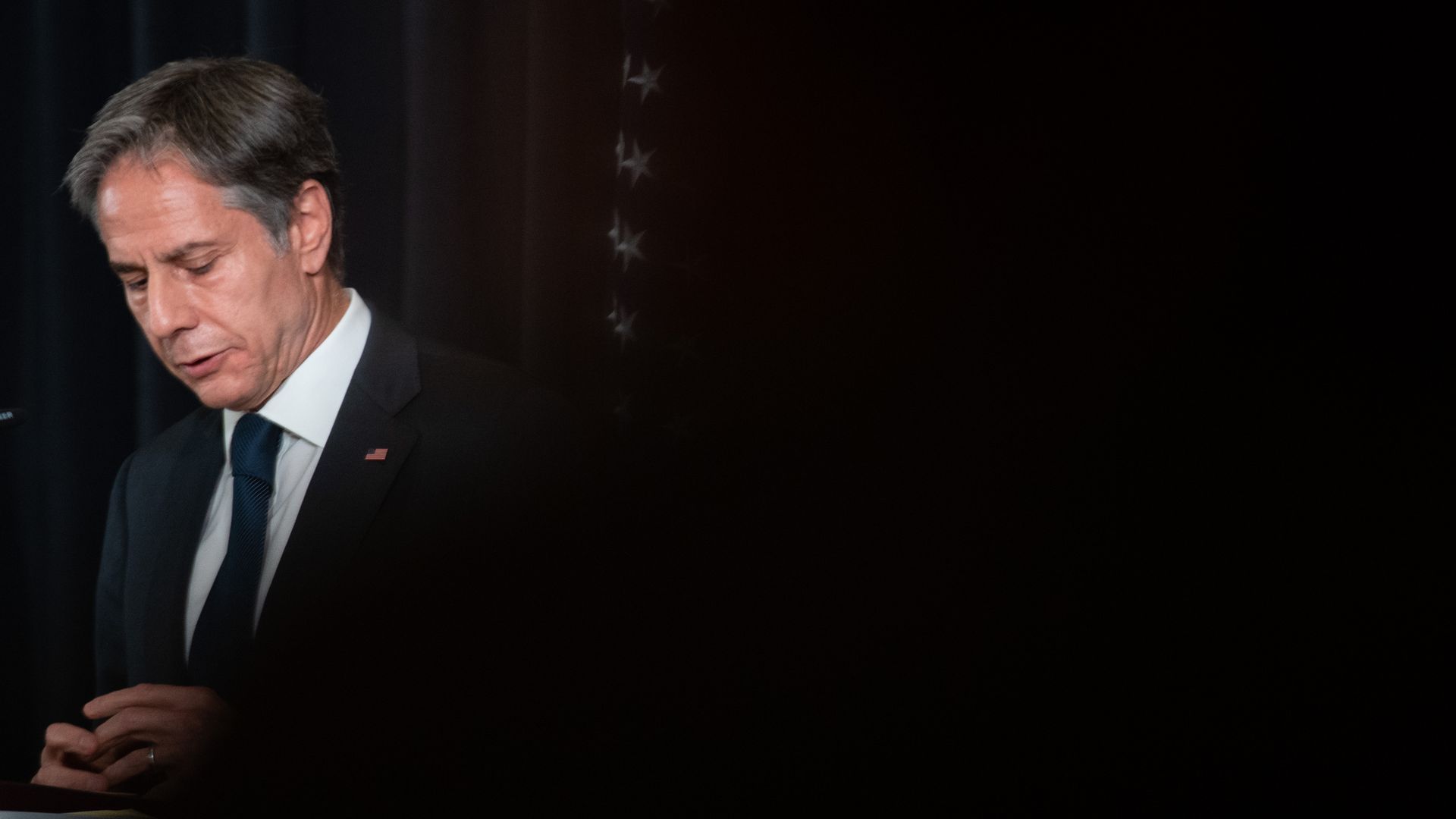 Secretary of State Tony Blinken looks down before speaking against a dark curtained background