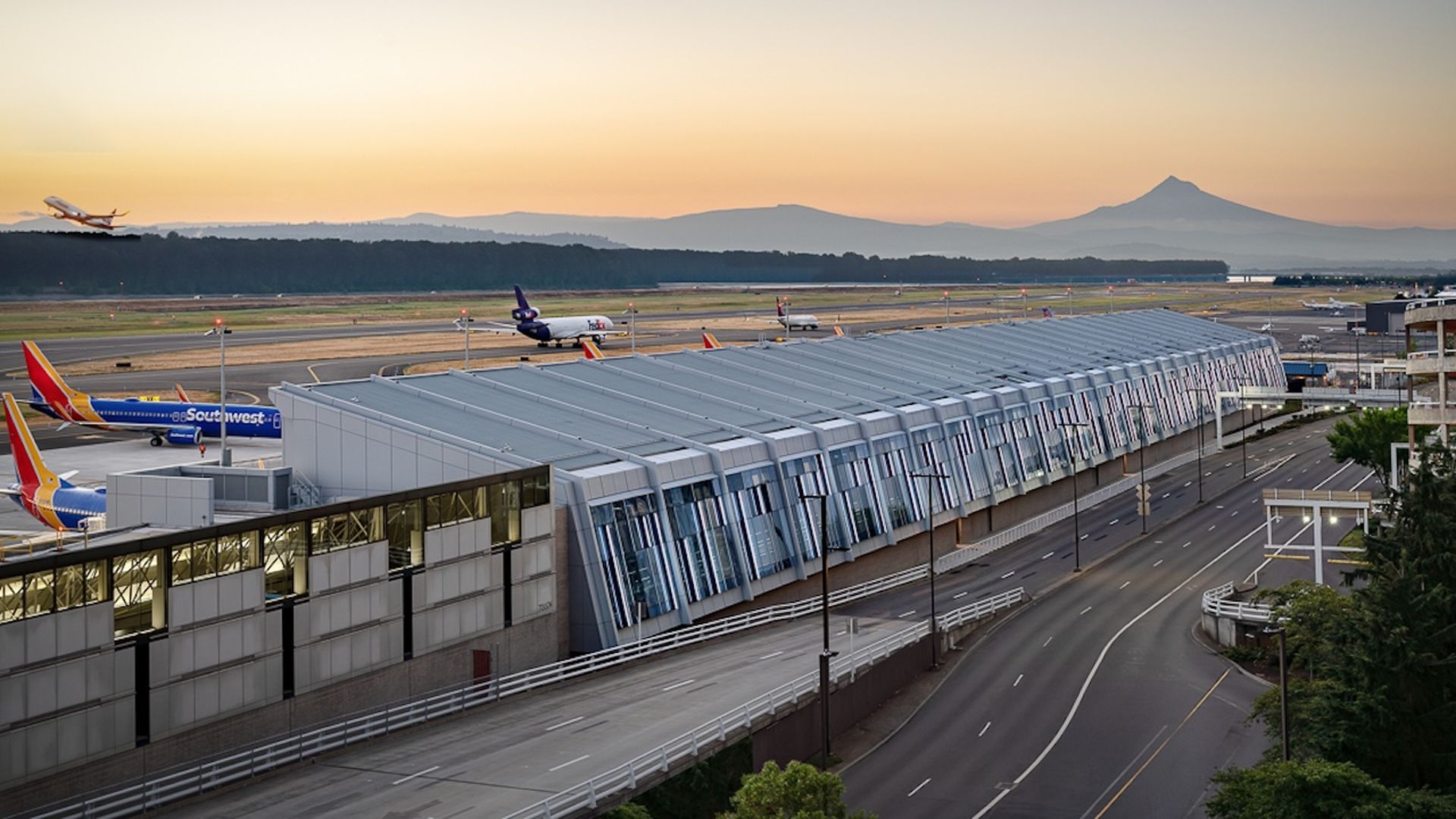 Airport terminal with Southwest Airlines planes, a FedEx plane, and a plane taking off at sunset, with mountains in the background and a clear sky.