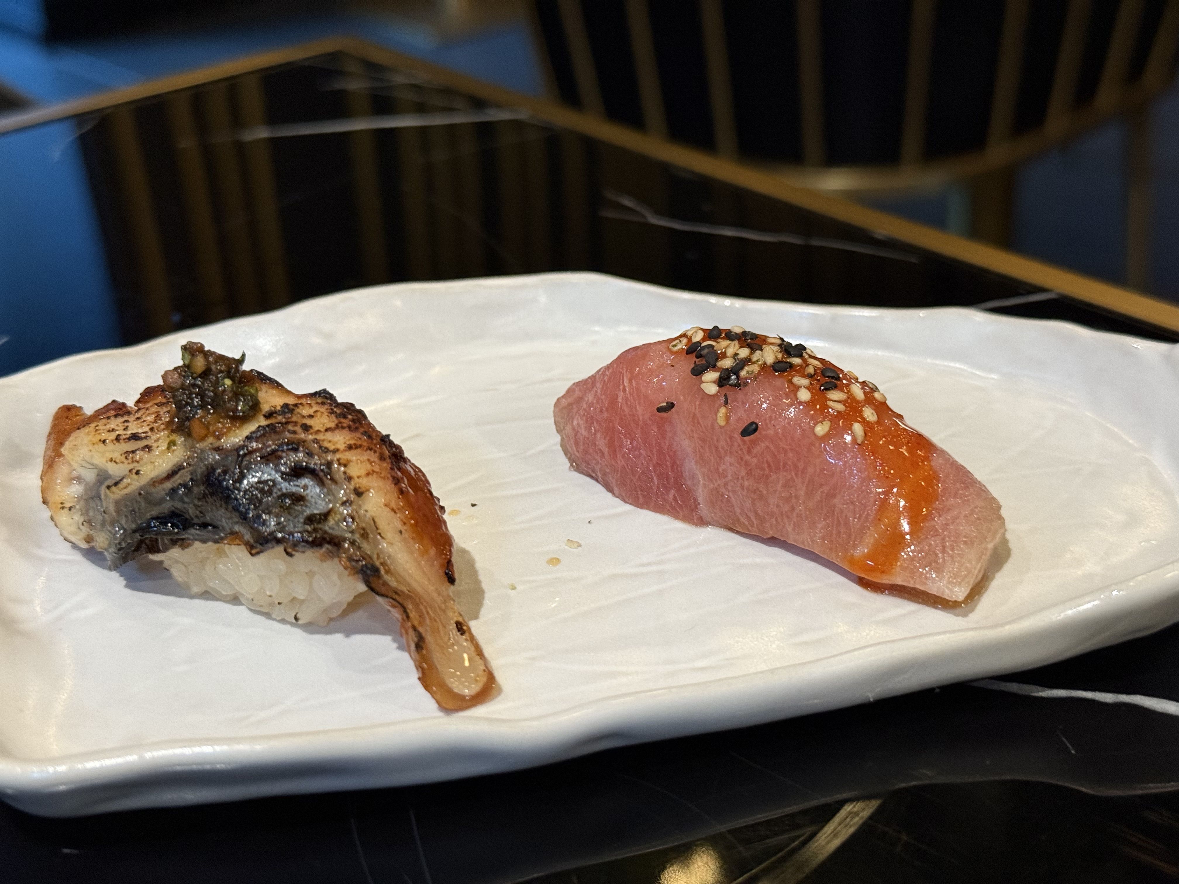 Two pieces of sushi on a white rectangular plate: one seared with dark sauce and garnish, the other pink tuna topped with sesame seeds and orange sauce, on a black table surface.