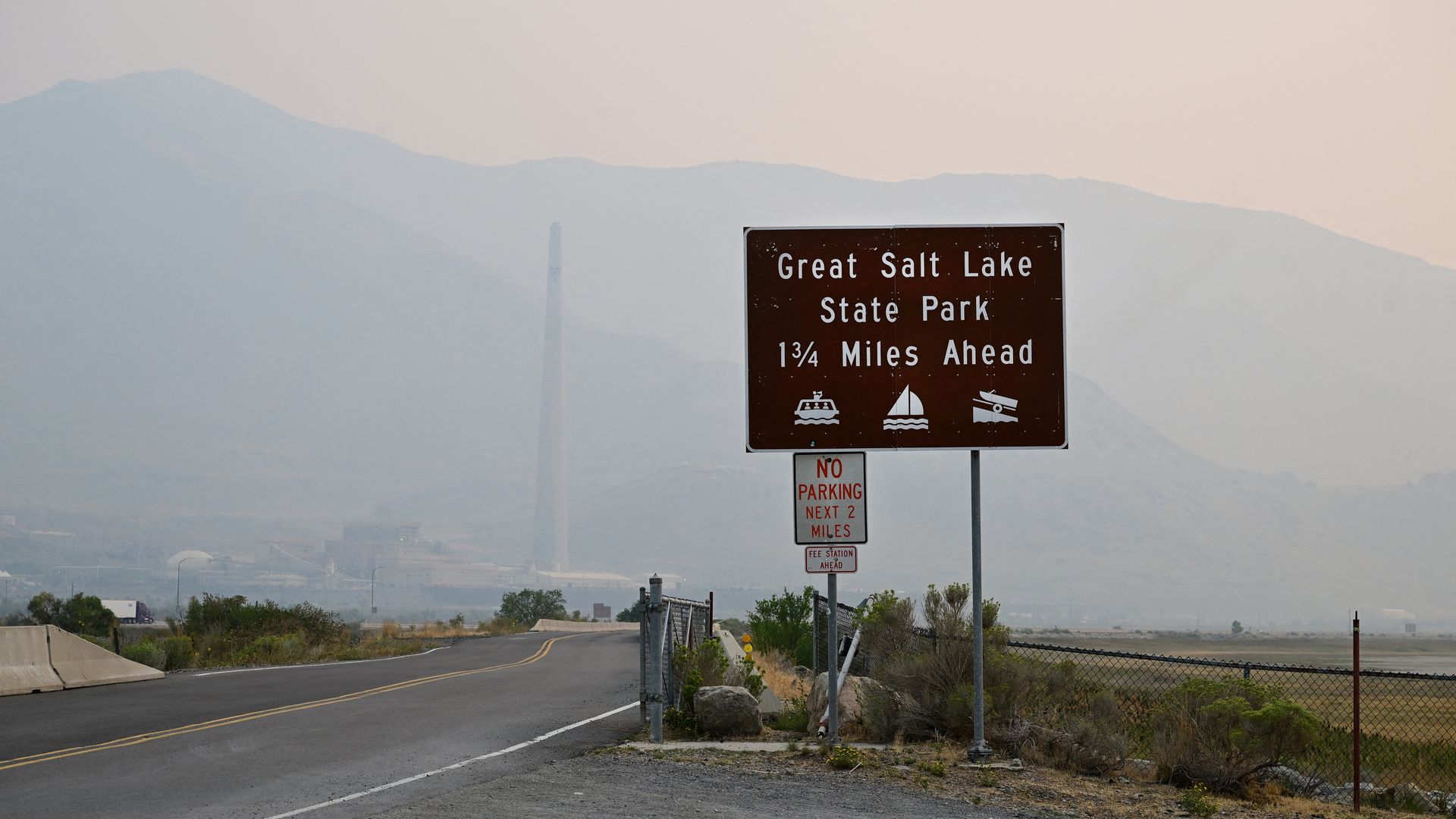 Haze obscures the mountains and buildings behind a sign that reads "Great Salt LAke State Park 1 3/4 miles ahead"