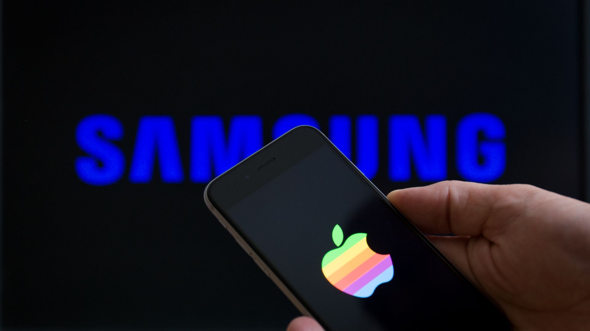 An iPhone with Apple logo on its screen under a Samsung logo sign