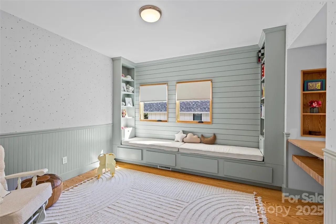 Bright room with light gray built-in bench under two windows, white cushions and star, cloud, and moon pillows, light wood floor, white rug, white rocking chair, and soft pale walls.