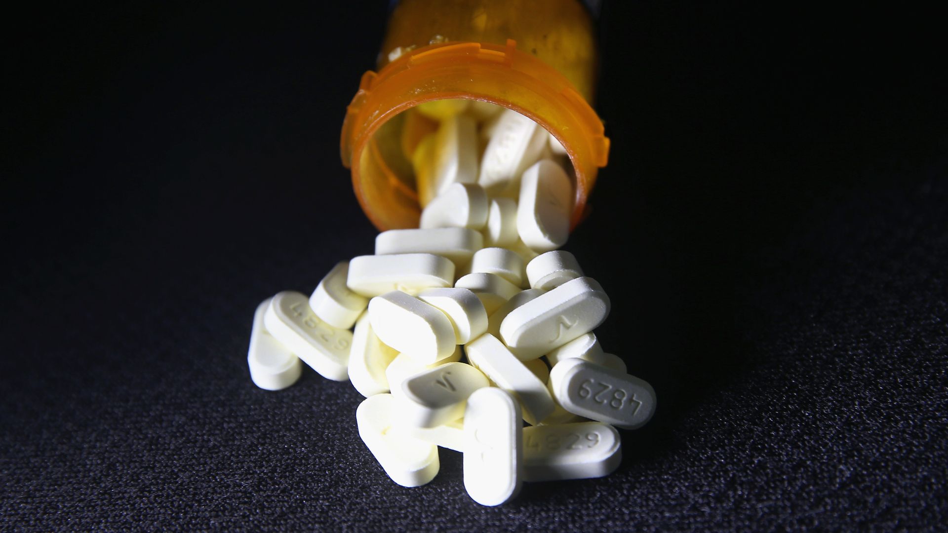 In this image, white pain pills fall out of an orange pill bottle.