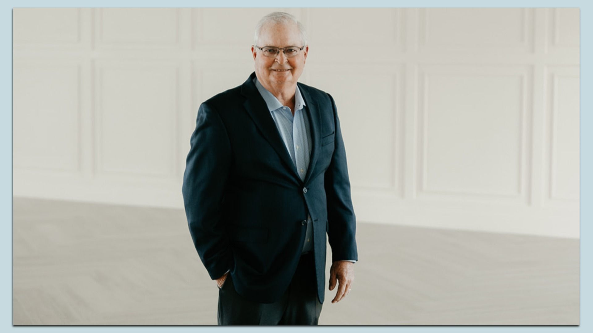 Dennis Albaugh with gray hair and glasses, wearing a dark suit and light blue shirt, stands in a bright, paneled white room.