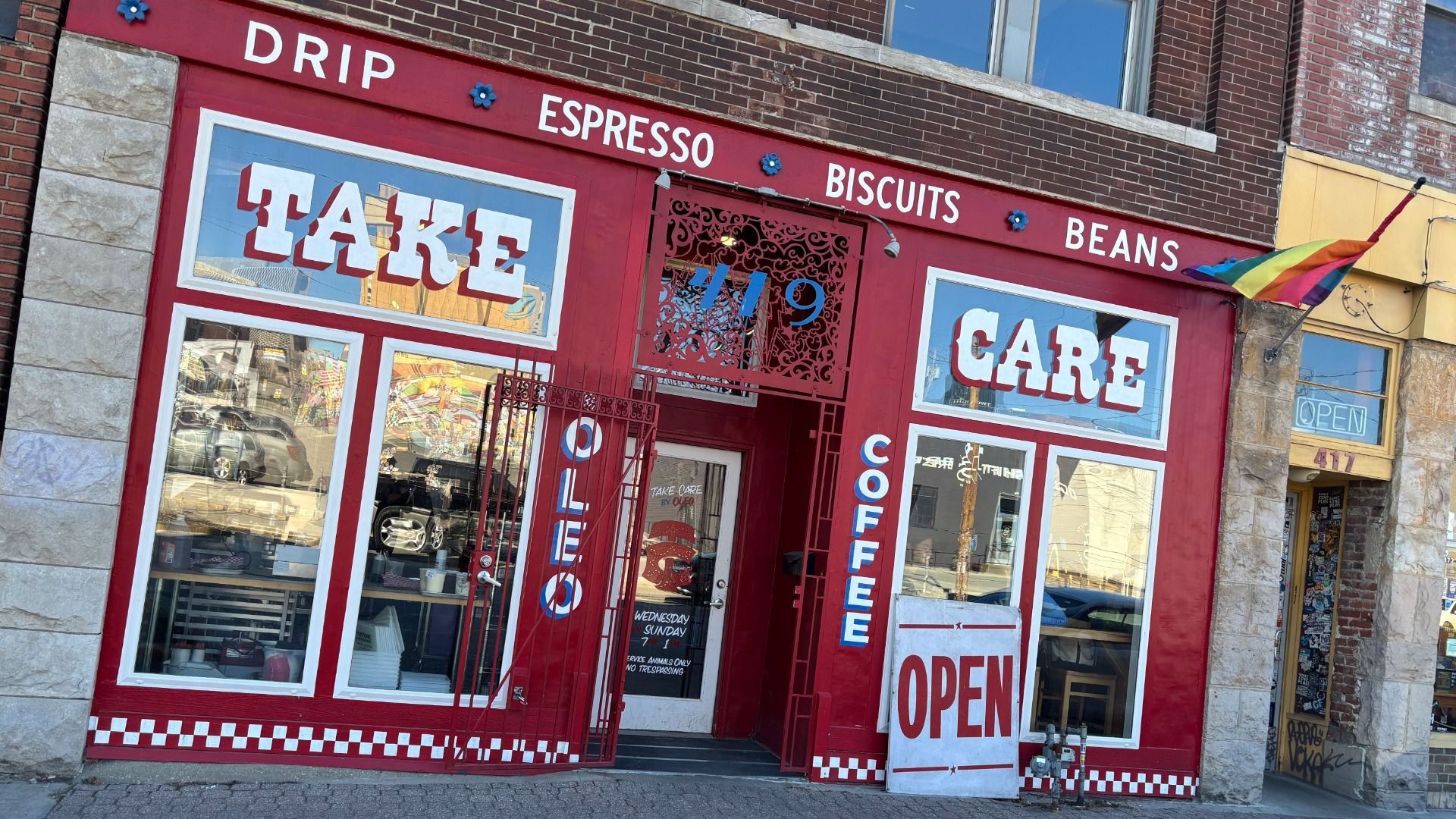 Red coffee shop storefront with white and blue lettering reading "TAKE CARE" and menu items like drip, espresso, biscuits, and beans. Rainbow pride flag flies on right side.
