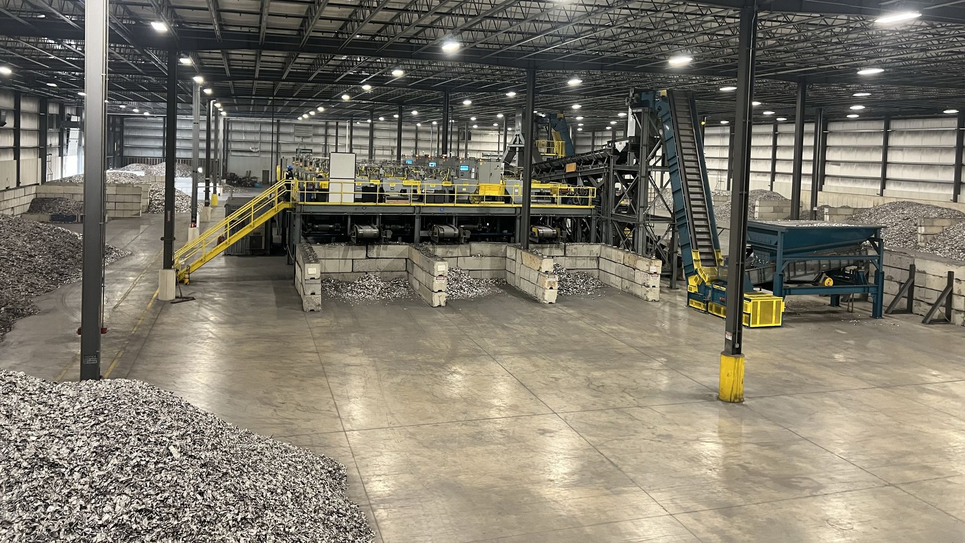 Large indoor recycling facility with piles of shredded metal, conveyor belts, sorting machinery, and yellow safety railings under high industrial lights in a spacious warehouse.