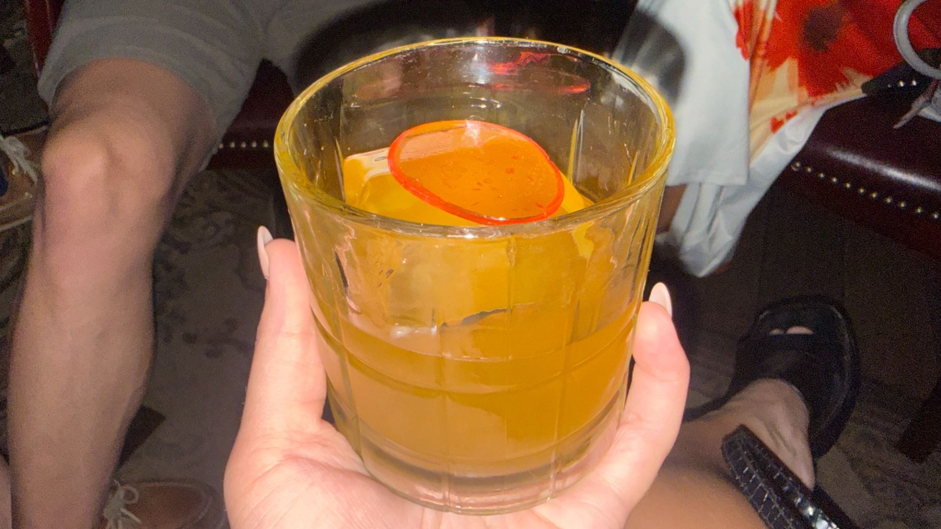 A clear glass tumbler of orange juice with ice, topped by a circular orange slice. A hand holds the glass; background shows a leg, sandals, and a colorful patterned shirt.