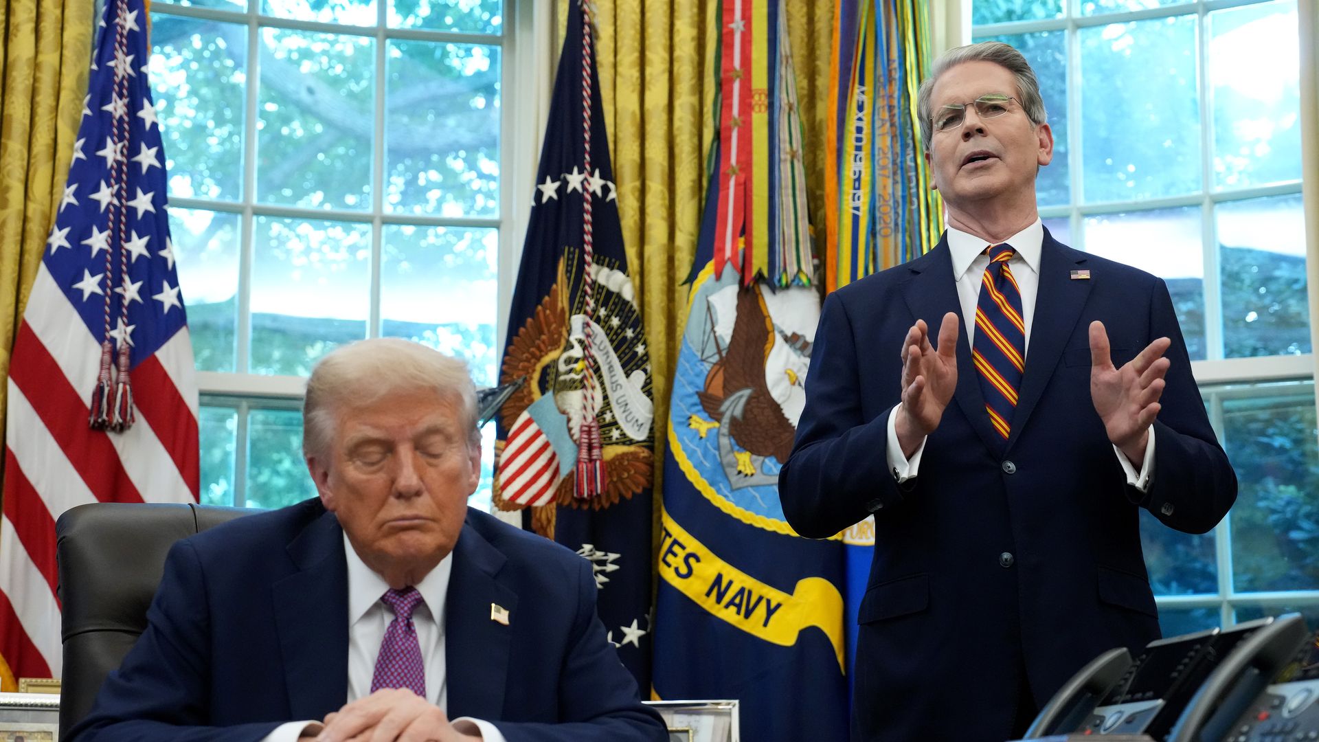 Treasury Secretary Scott Bessent stands, gesturing, next to a seated President Trump in the Oval Office. 