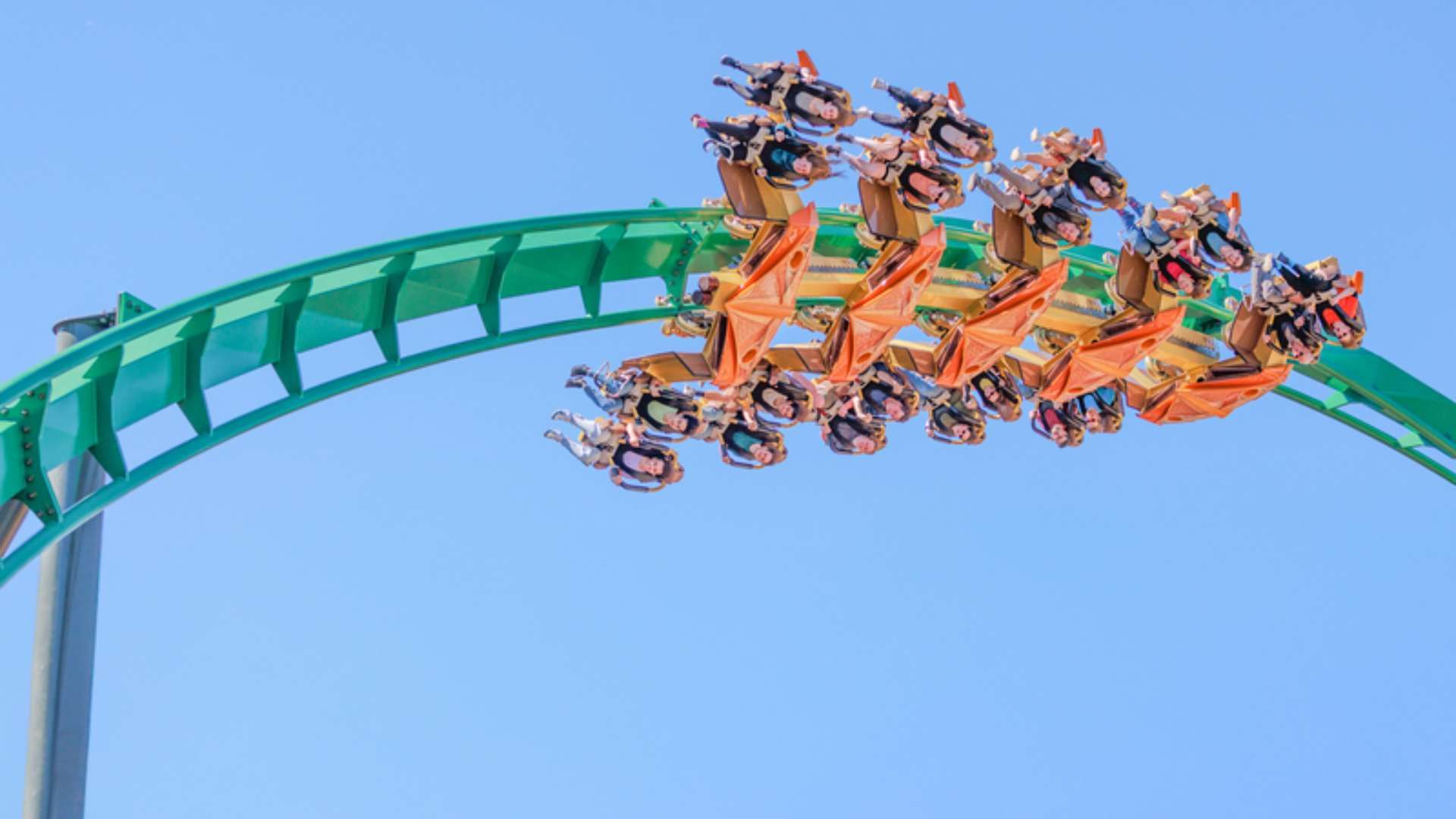 Bright blue sky above a green roller coaster arc; an orange train carries numerous riders in colorful clothing as they zoom along the track.