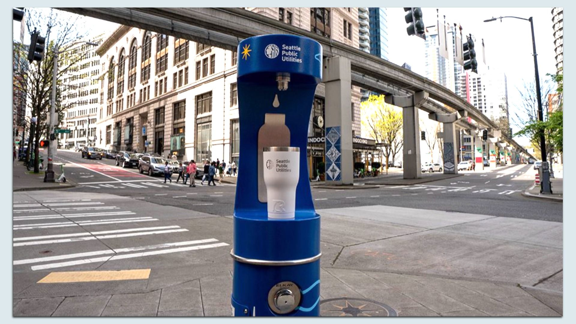 A blue Seattle Public Utilities drinking fountain in the foreground on a city street, with an elevated track, tall buildings, pedestrians crossing a crosswalk, and cars along the curb.