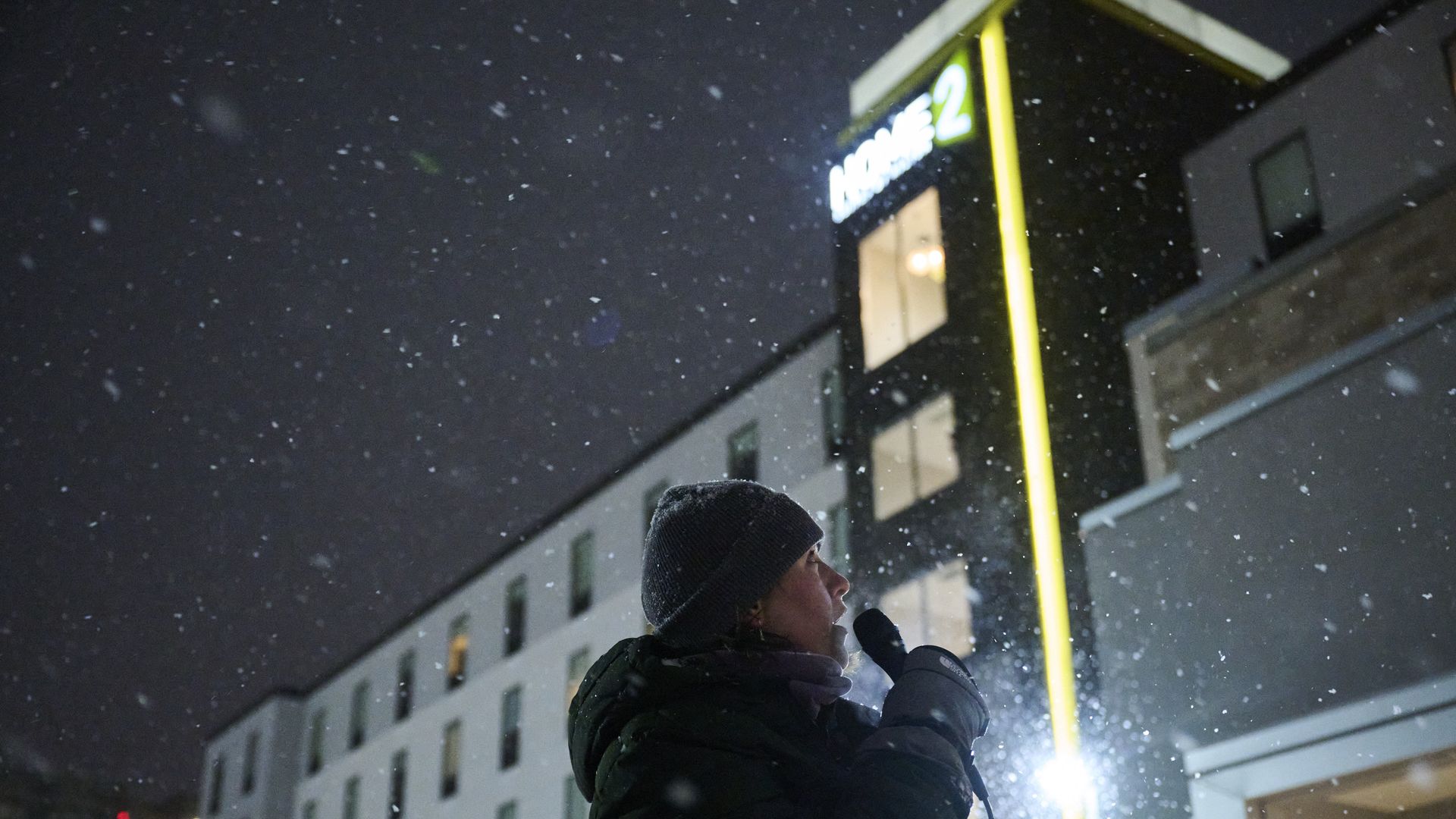 A woman stands outside of a hotel holding a microphones. The hotel has a green Home 2 sign. 