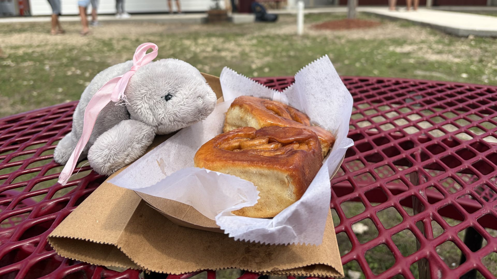 A soft gray plush animal with a pink ribbon rests on a red metal picnic table beside a golden-brown pastry wrapped in white paper.