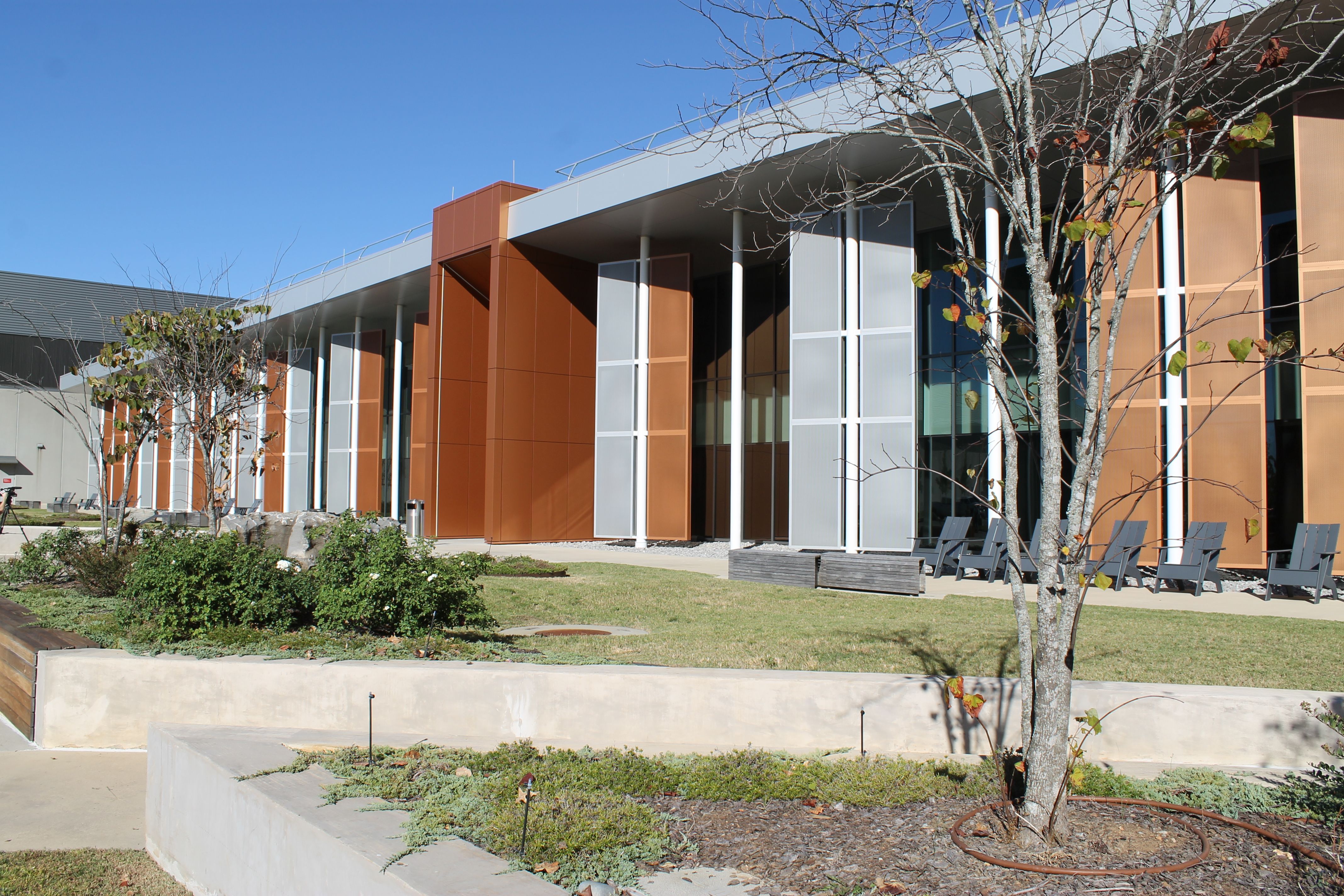 Modern building with orange-brown panels and large glass windows, white columns under a covered walkway; bare trees, blue sky, and outdoor chairs along a landscaped front area.