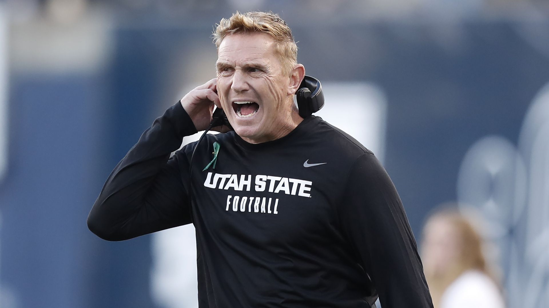 A man wearing a black shirt with the words "Utah State Football" yells while removing a headset.