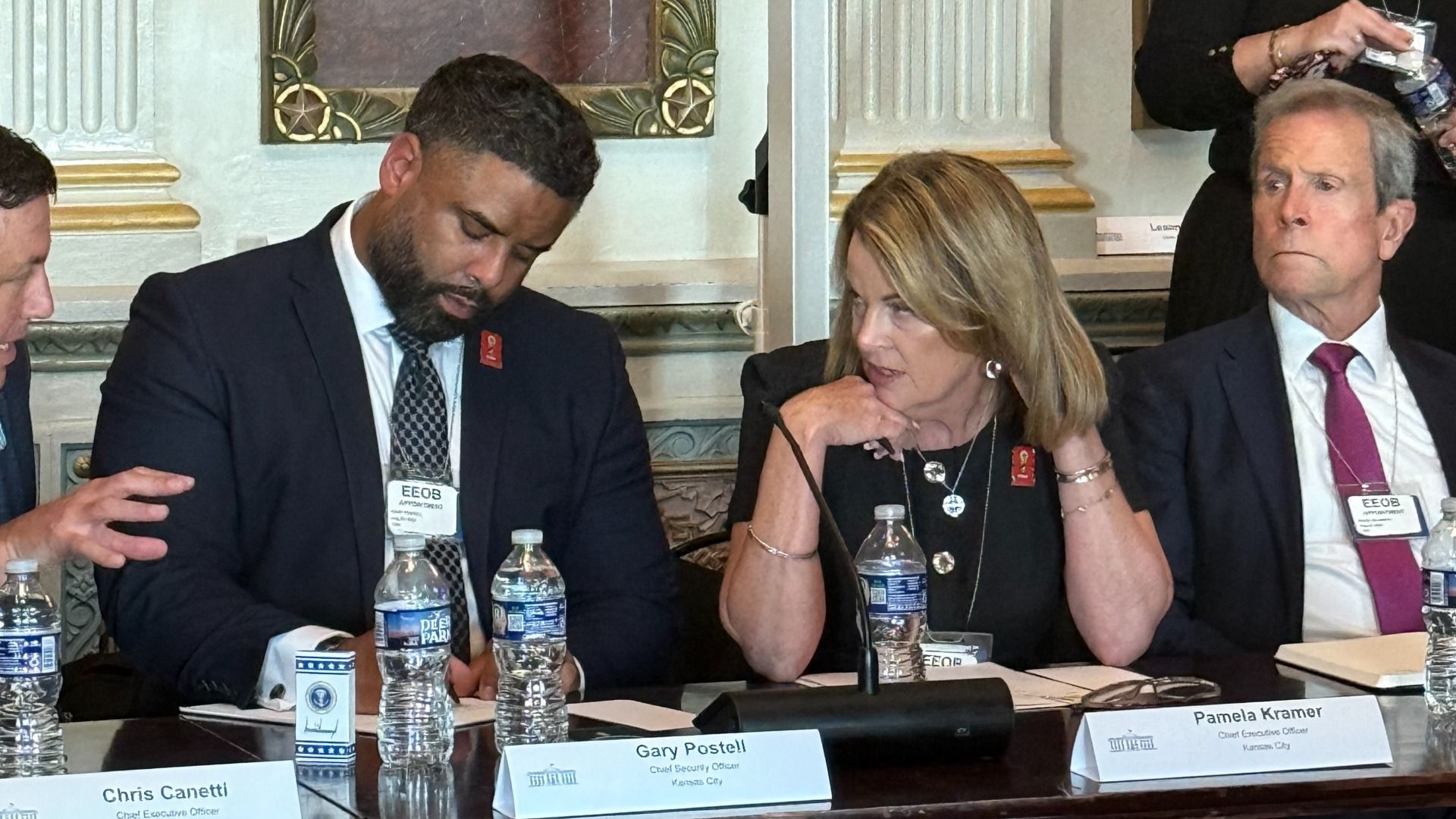 Three people in business attire seated at a conference table with nameplates and water bottles, including Gary Postell and Pamela Kramer, in a formal room with ornate decor.