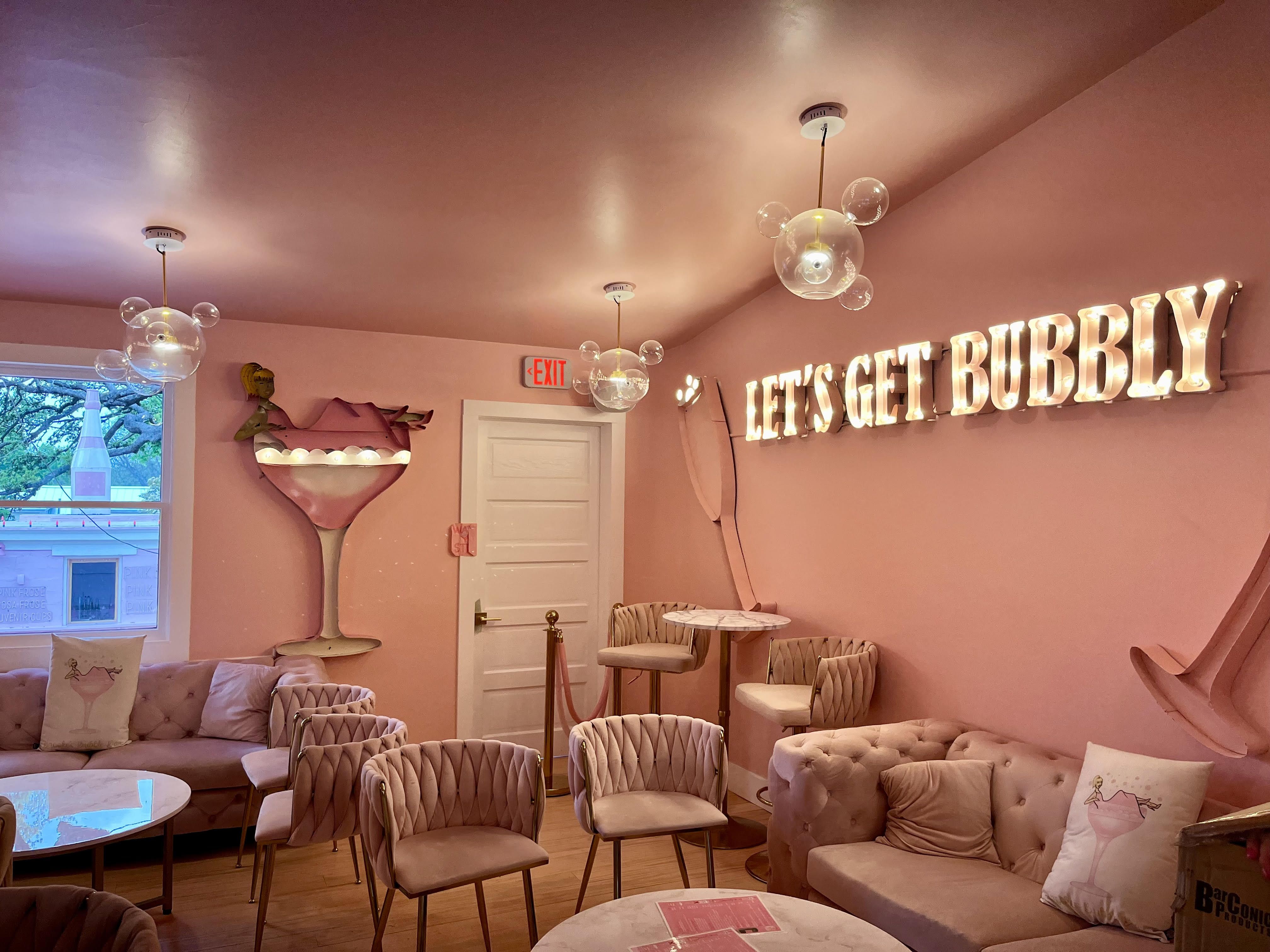 An all-pink room with cushy pink chairs and a couch with pink pillows. Lit-up wall text reads "Let's Get Bubbly."