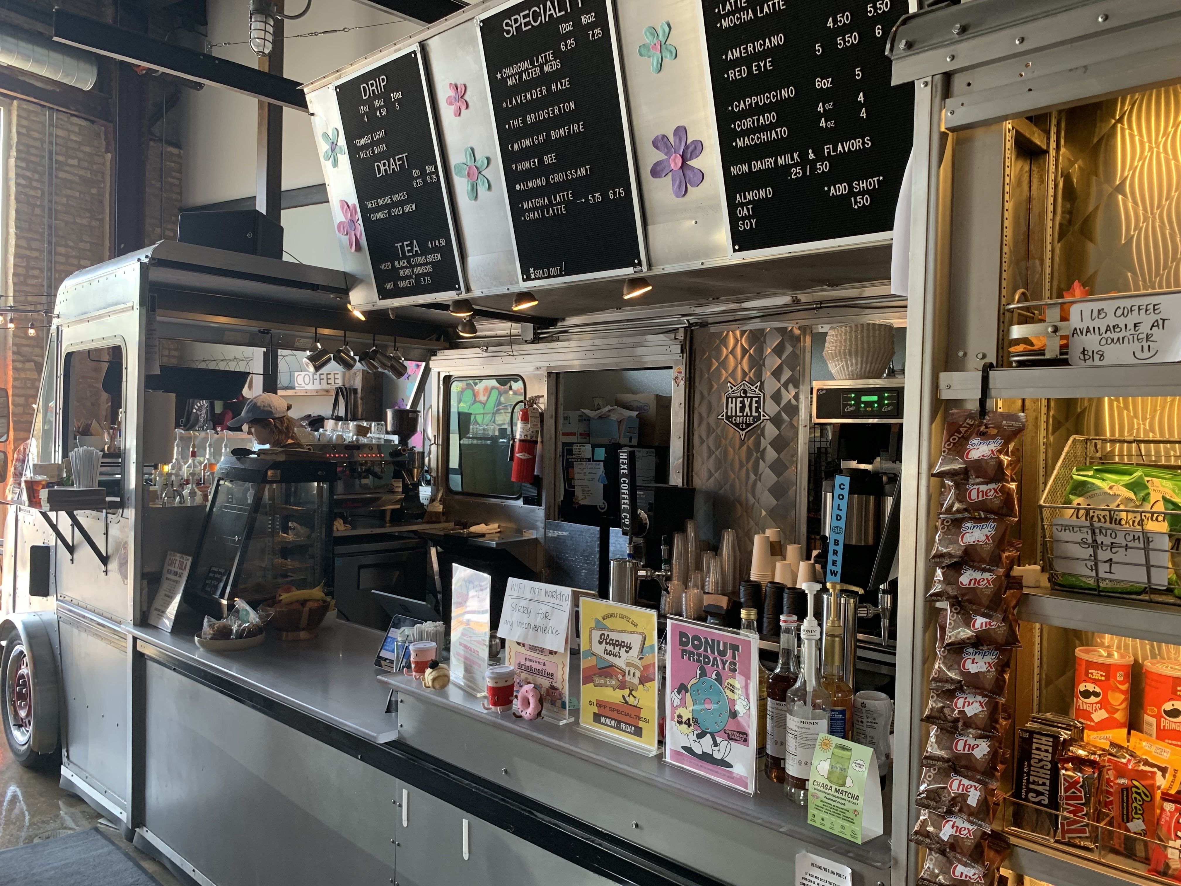 Interior of a coffee shop with a metal serving counter, hanging black menu boards, coffee syrups, snacks like Pringles and Hershey's, and a small barista preparing drinks.