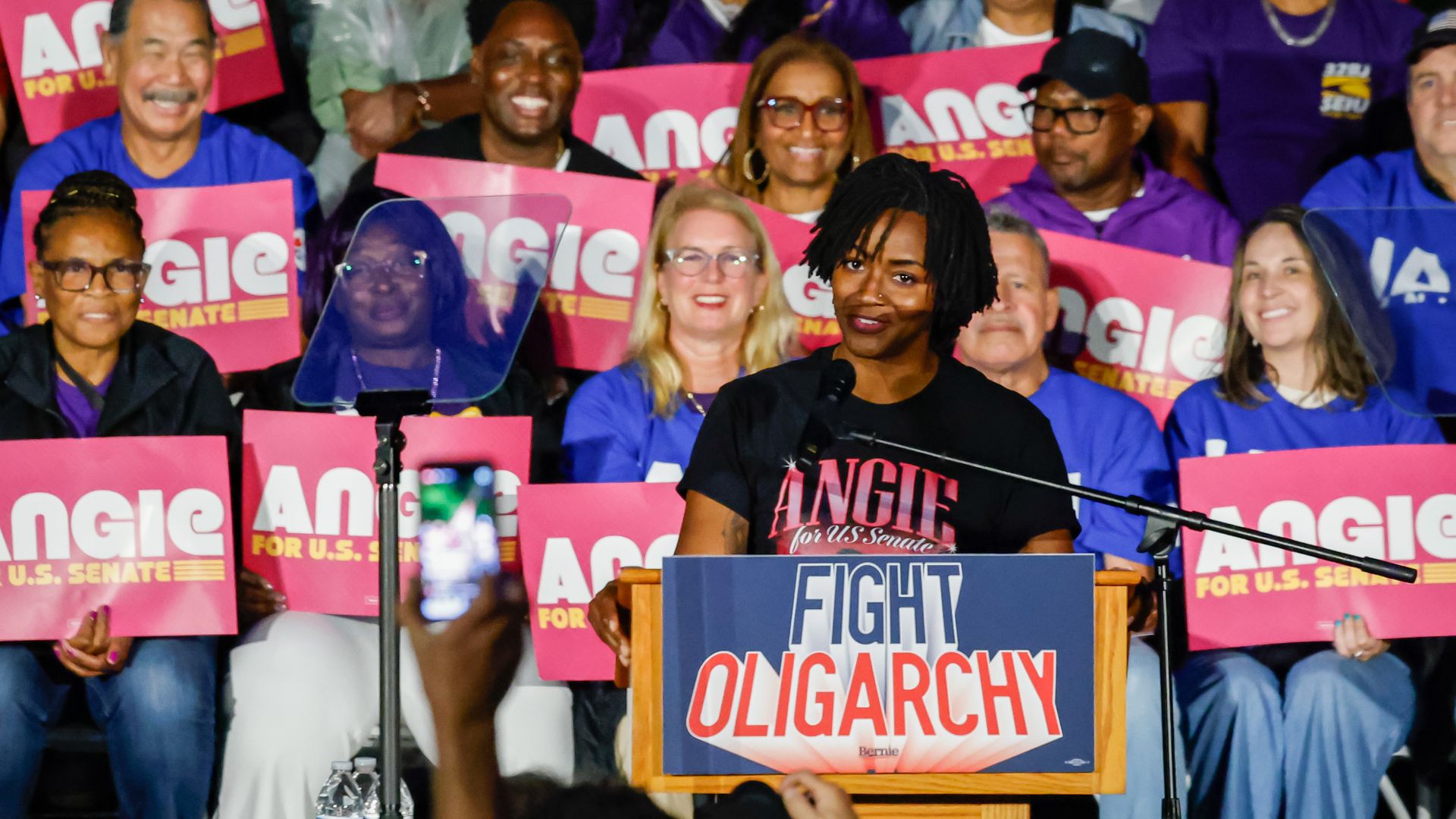 Angie Nixon speaks at a podium displaying a "Fight Oligarchy" sign, surrounded by supporters holding signs promoting her U.S. Senate campaign.