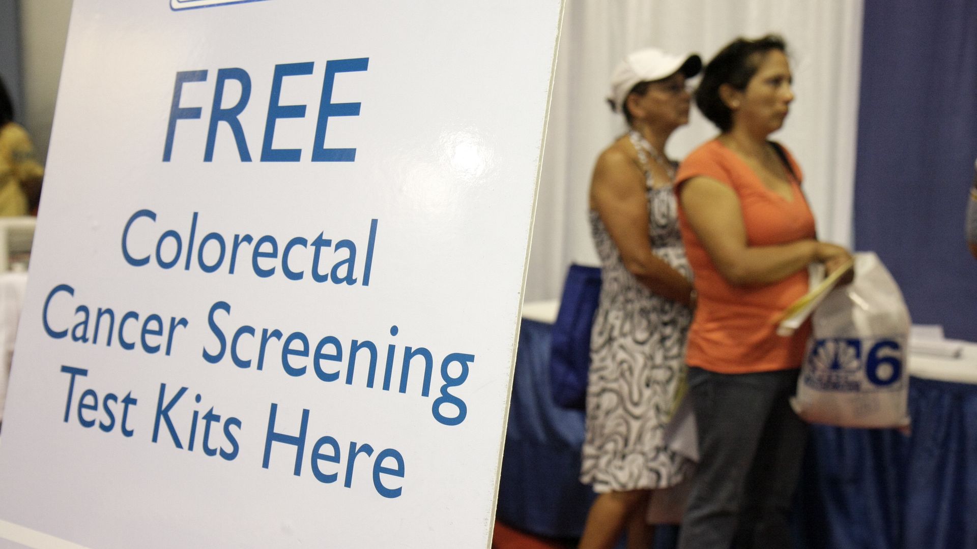 Women wait in line near a sign advertising free colorectal cancer screening test kits.