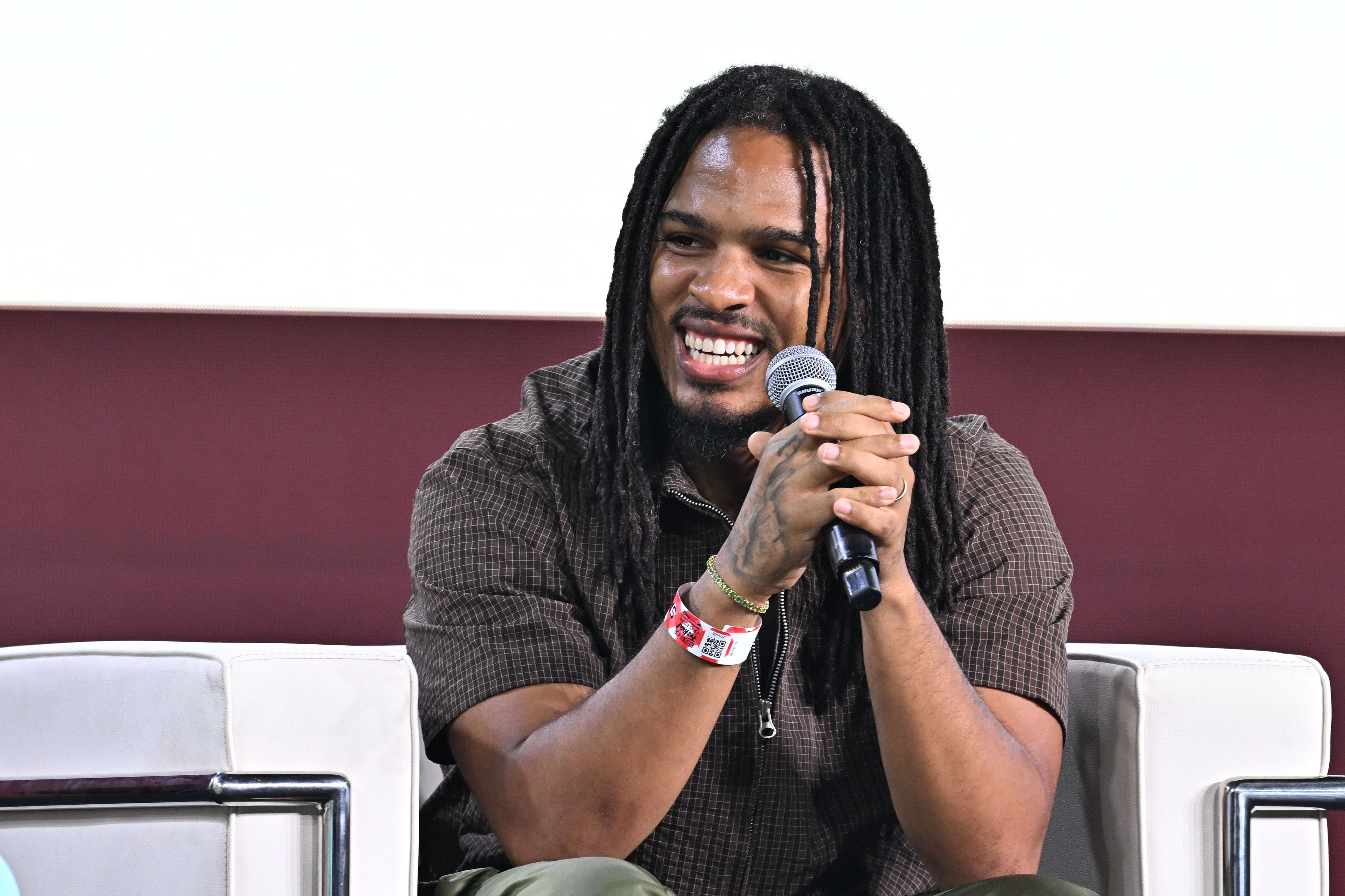 Smiling man with long dreadlocks wearing a brown checkered shirt, holding a microphone with tattooed hands and wristbands, sitting on a white chair against a maroon and white backdrop.