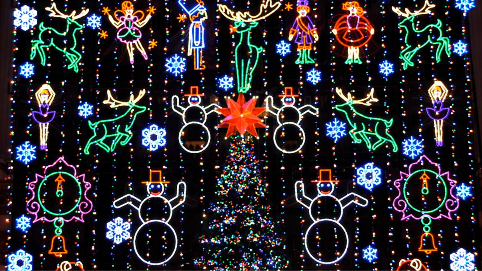 Colorful Christmas light display with snowmen, reindeer, ballet dancers, toy soldiers, clocks, snowflakes, and a decorated Christmas tree topped with a red star.