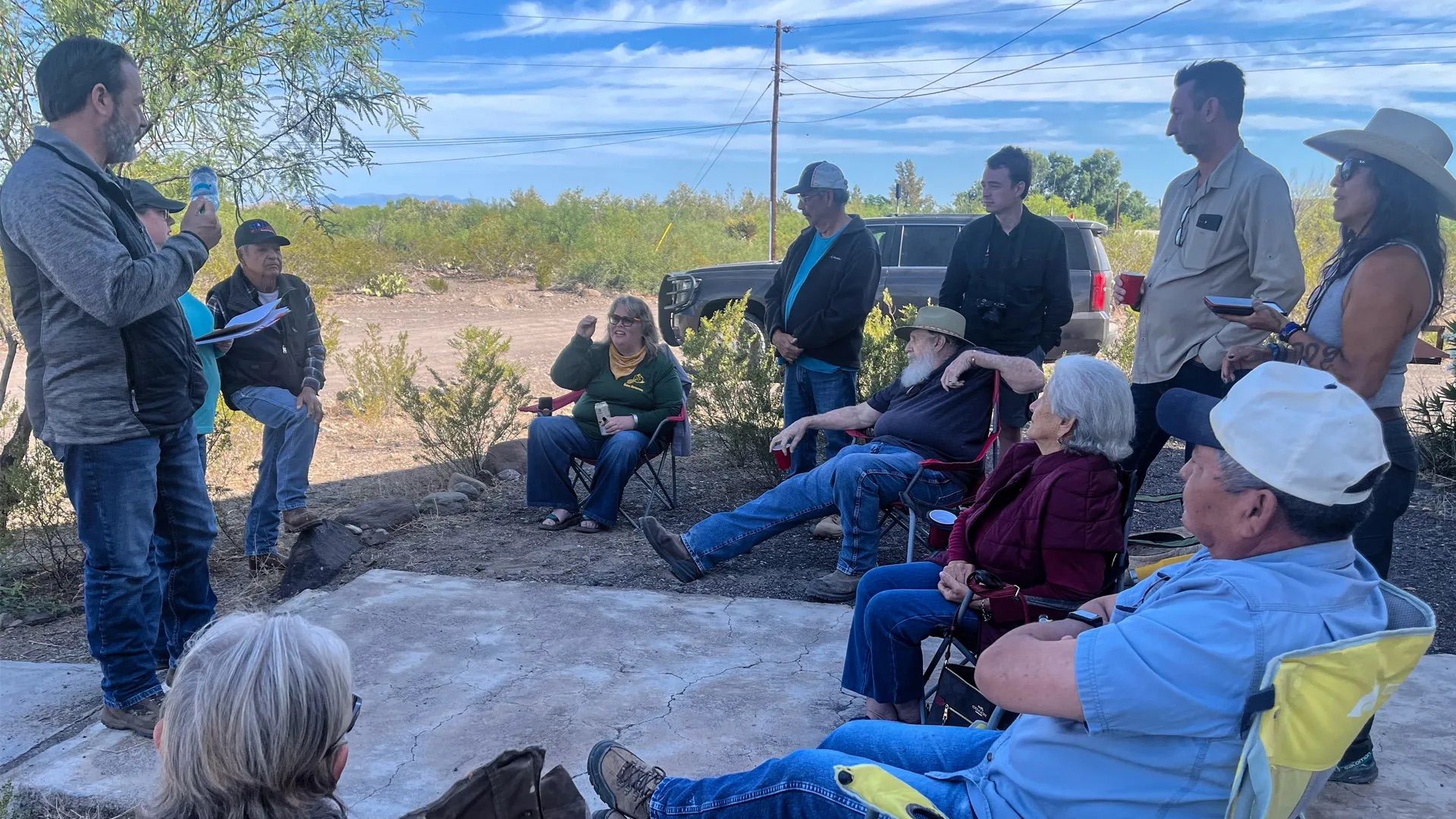 An outdoor gathering in the Big Bend desert in Texas: people seated and standing in a circle as a man on the left speaks, holding a water bottle and papers; a gray SUV and power lines are behind them.