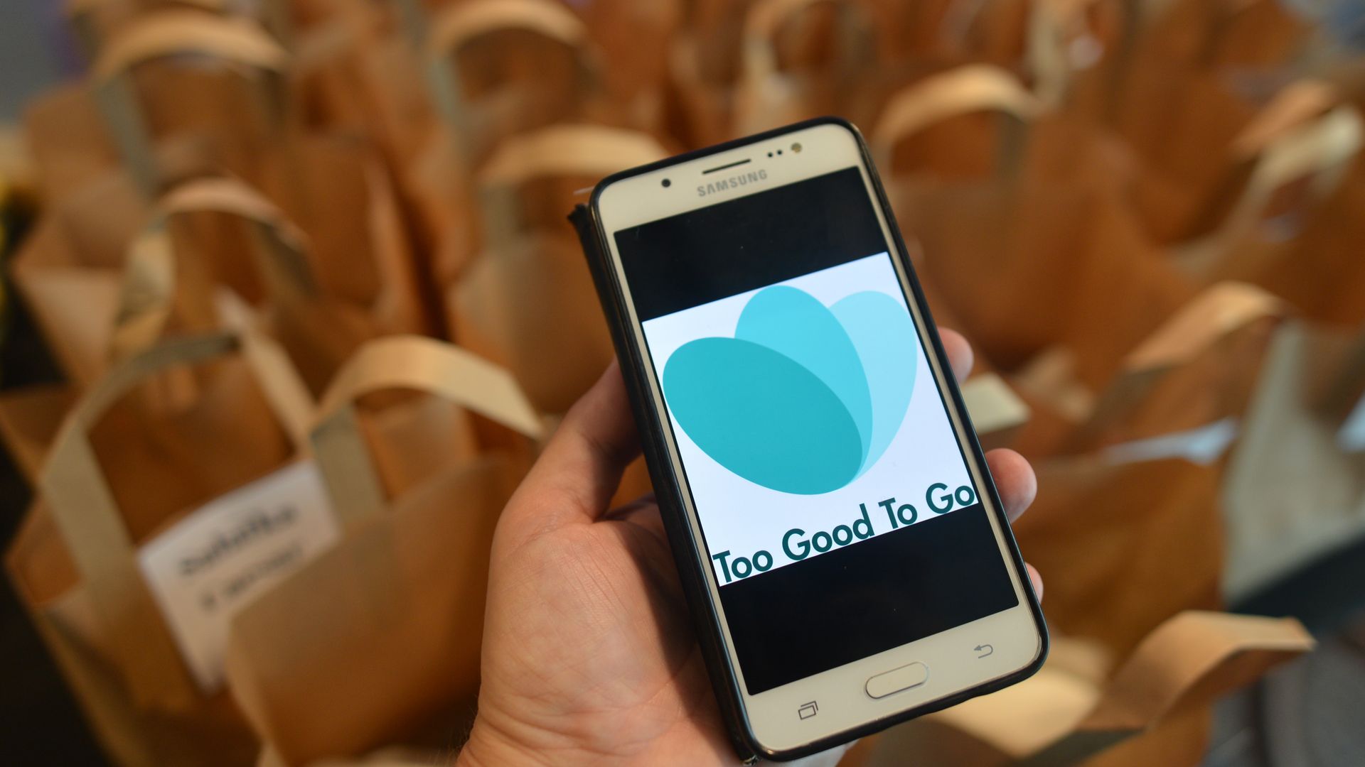 A photo of someone holding a cell phone with the too good to go logo with lots of paper bags in the background
