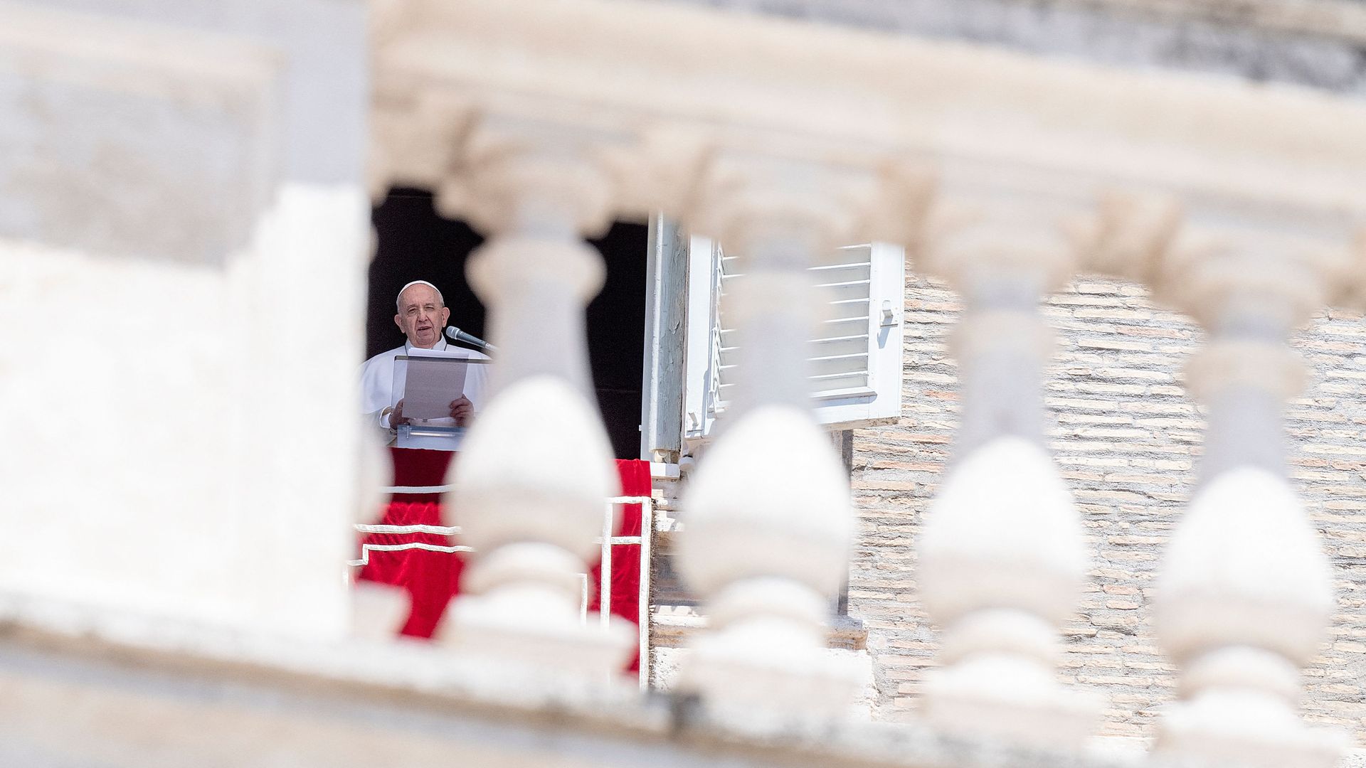 Photo of Pope Francis wearing white and speaking into a mic as he holds a piece of paper, juxtaposed against columns